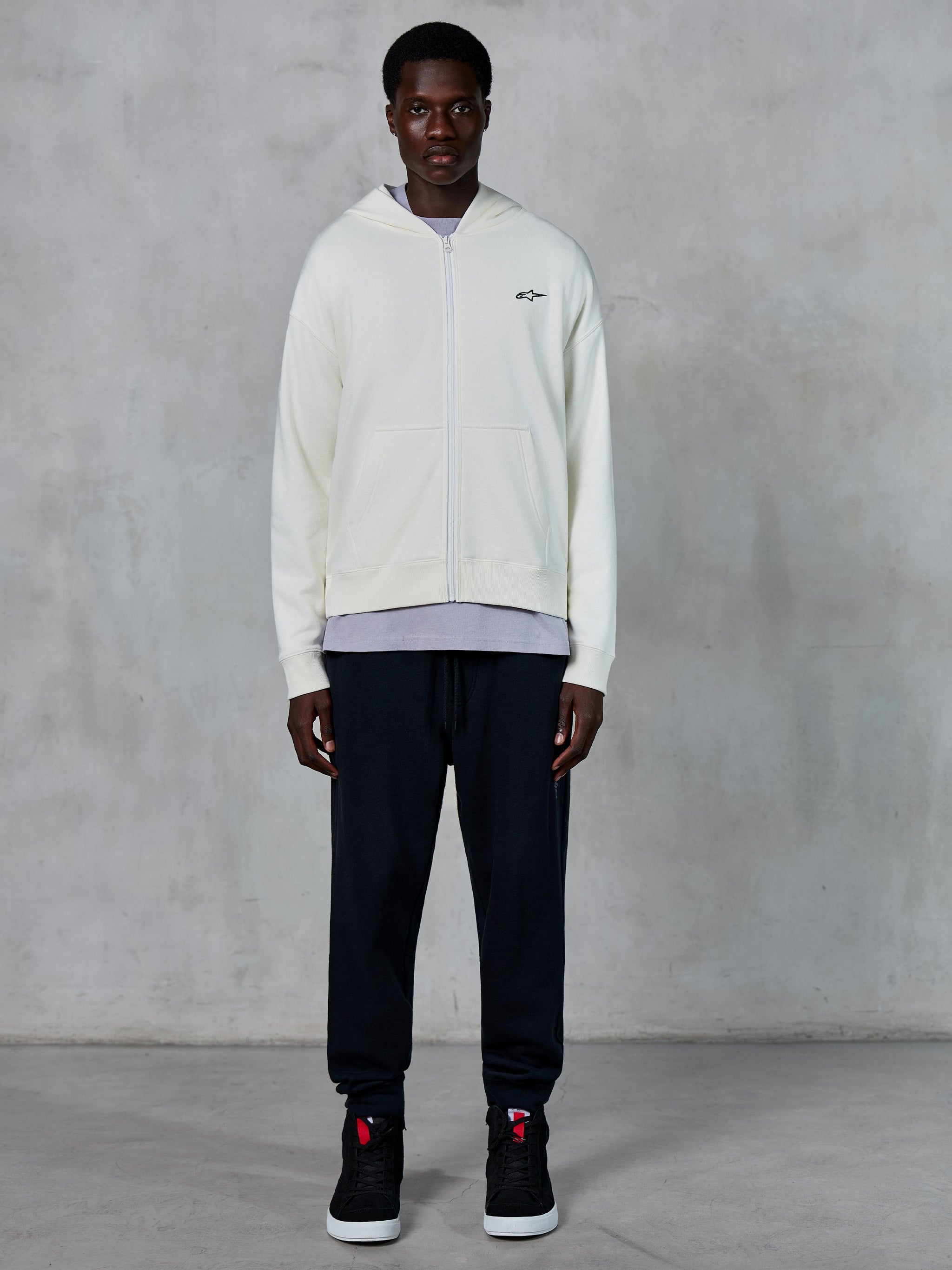 Alpinestars Essentials Ageless Zip Hoodie, off-white hoodie met volledige rits, ontspannen pasvorm met een klein zwart logo op de borst, gedragen met een zwarte joggingbroek en sneakers, casual motorsport-geïnspireerde streetwear.