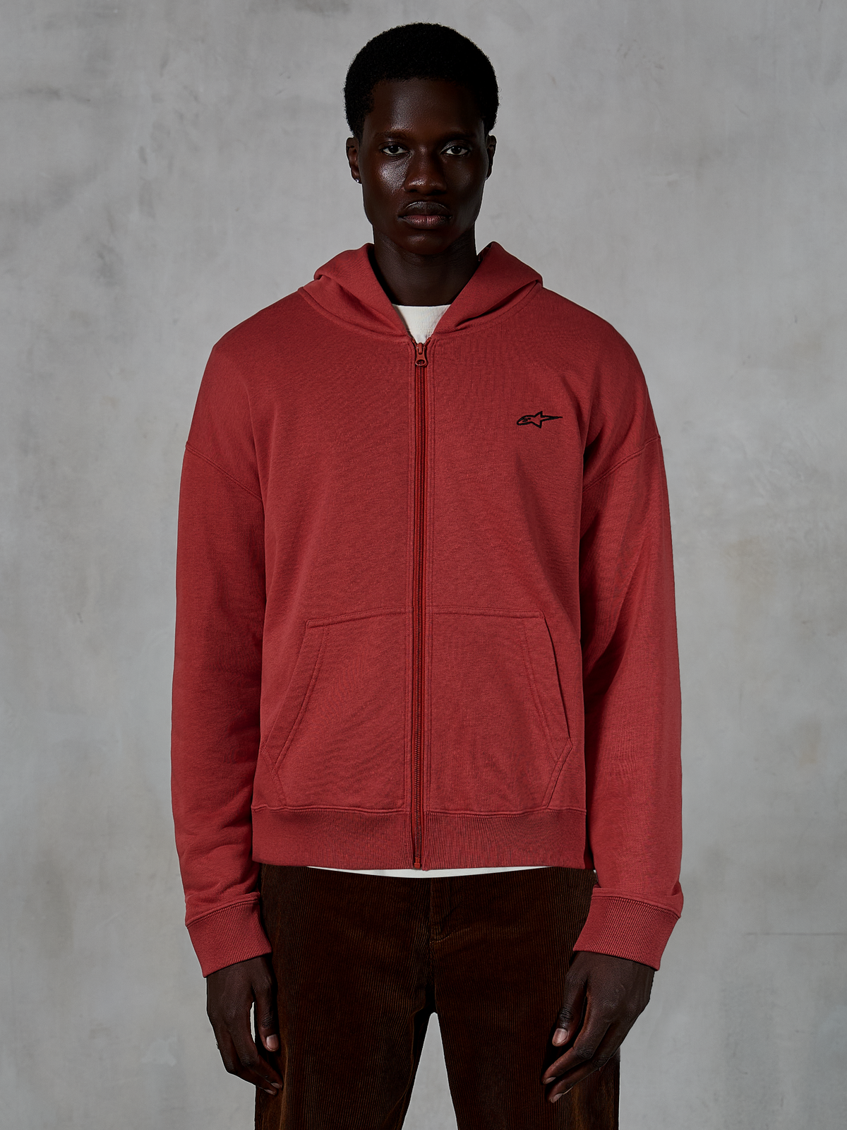 Alpinestars Essentials Ageless Zip Hoodie, steenrode hoodie met volledige ritssluiting, gedragen door model met bruine corduroy broek, voorzien van een klein zwart logo op de borst, ontspannen pasvorm casual streetwear