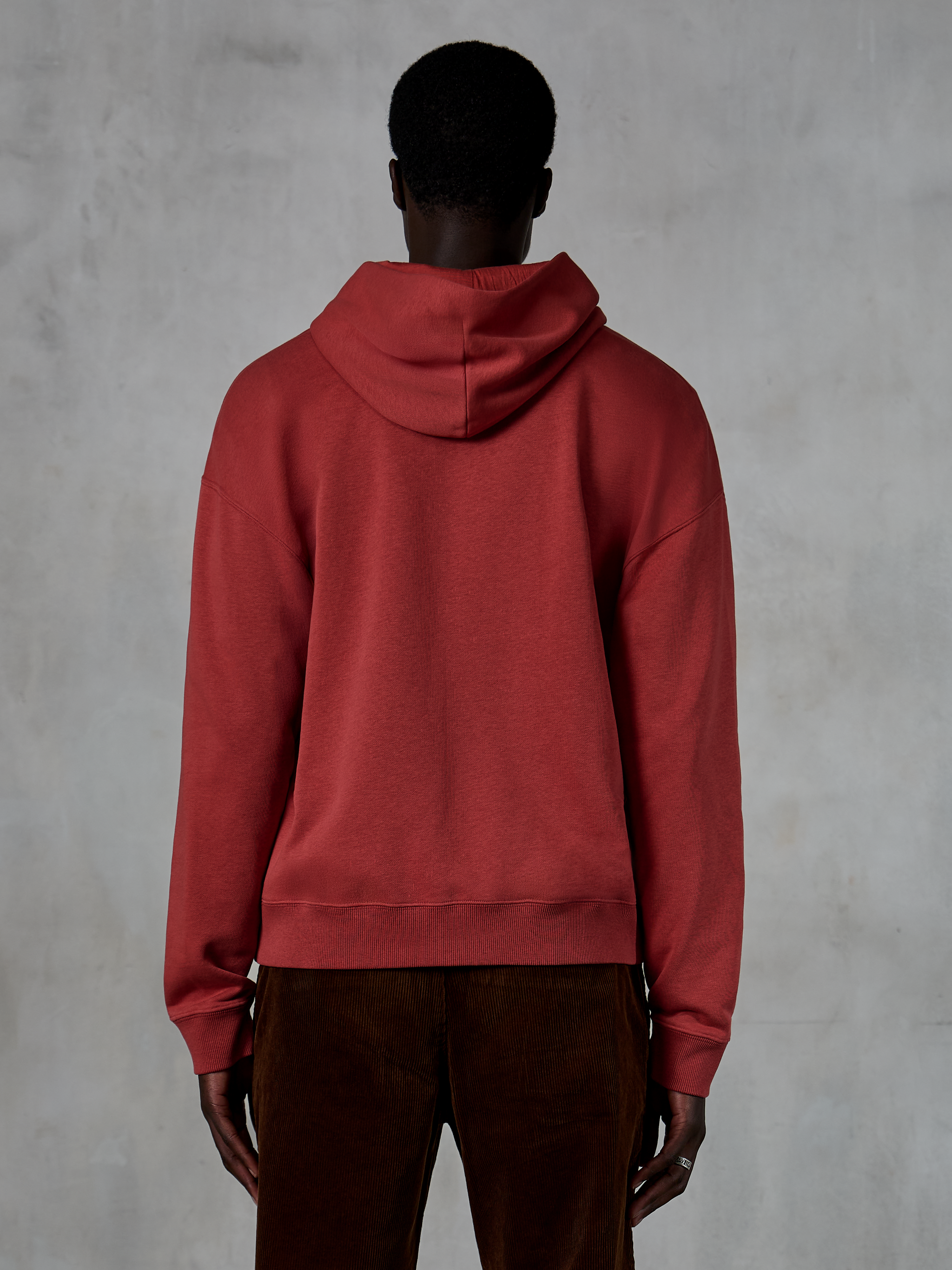 Alpinestars Essentials Ageless Zip Hoodie, Brick red, back view showing relaxed fit and drawstring hood, worn with brown corduroy pants, casual motorsport-inspired streetwear