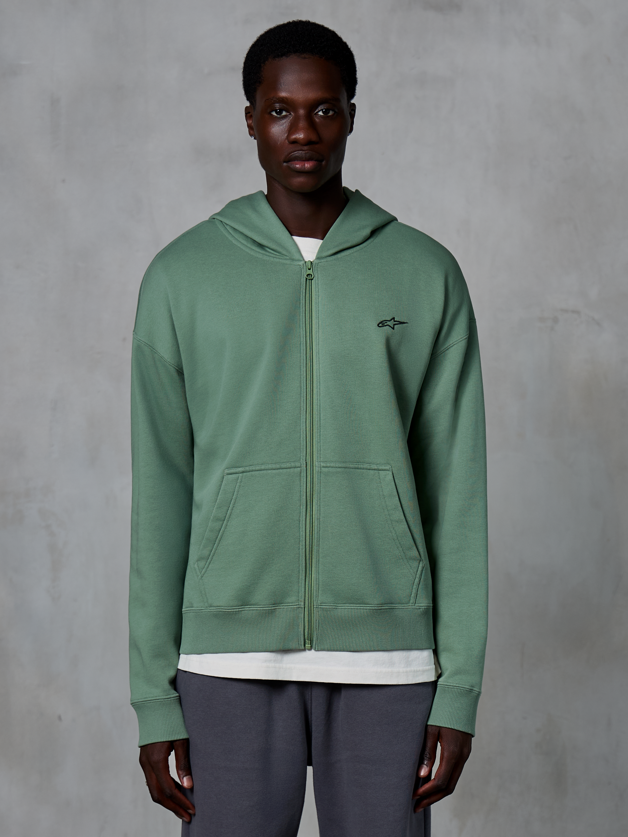 Alpinestars Essentials Ageless Zip Hoodie, casual streetwear hoodie met rits, Cactus groen, voorzien van een subtiel geborduurd logo op de borst, gedeelde kangoeroezakken en geribbelde manchetten, gedragen door een mannelijk model tegen een grijze achtergrond met textuur