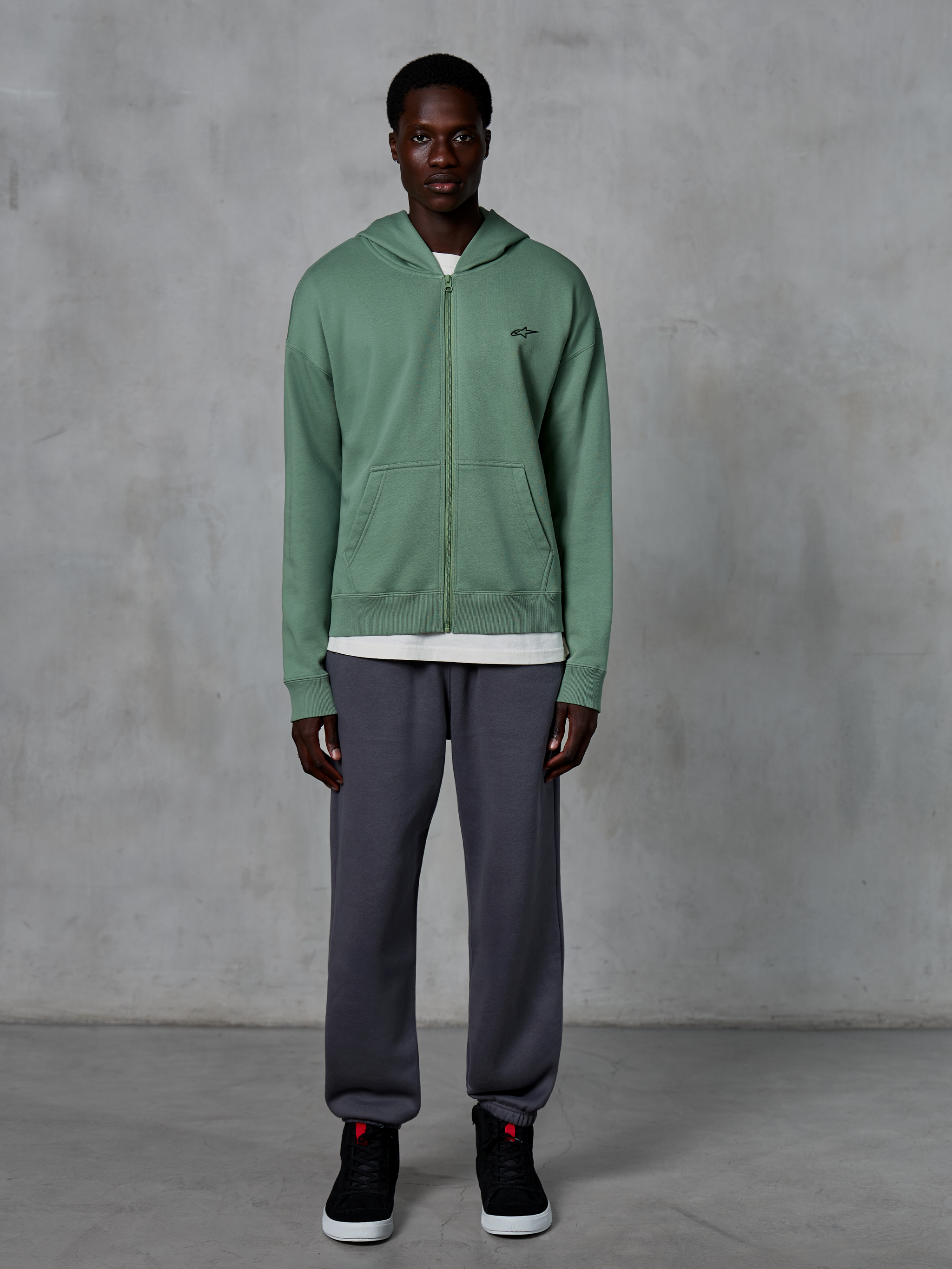 Alpinestars Essentials Ageless Zip Hoodie, hoodie met rits, Cactus Green, gedragen door een model met donkergrijze joggers en zwarte sneakers, klein zwart Alpinestars-logo op de borst, ontspannen pasvorm, casual streetwear