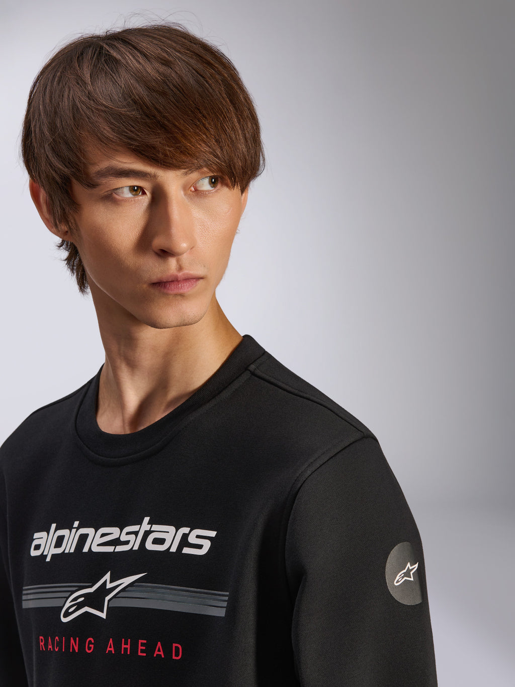 Alpinestars Bettern Crew sweatshirt, zwart, met wit Alpinestars-logo, grijze strepen en rode 'RACING AHEAD'-tekst, rond sterlogo op de mouw, casual streetwear geïnspireerd op de motorsport