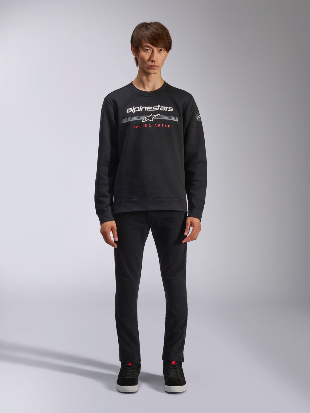 Alpinestars Bettern Crew, zwarte sweatshirt met ronde hals, wit Alpinestars-logo met grijze horizontale strepen en rode 'RACING AHEAD' tekst, casual motorsport streetwear, gedragen door model met zwarte broek en sneakers