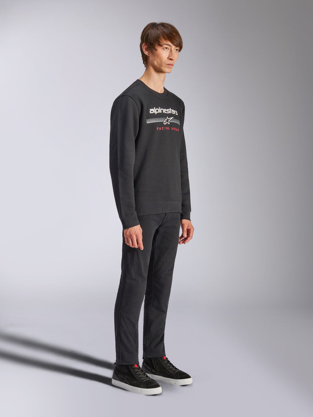 Alpinestars Bettern Crew Sweatshirt, zwarte pullover met ronde hals, wit Alpinestars-logo met 'RACING AHEAD' in het rood op de borst, gecombineerd met een zwarte broek en sneakers, casual op motorsport geïnspireerde streetwear