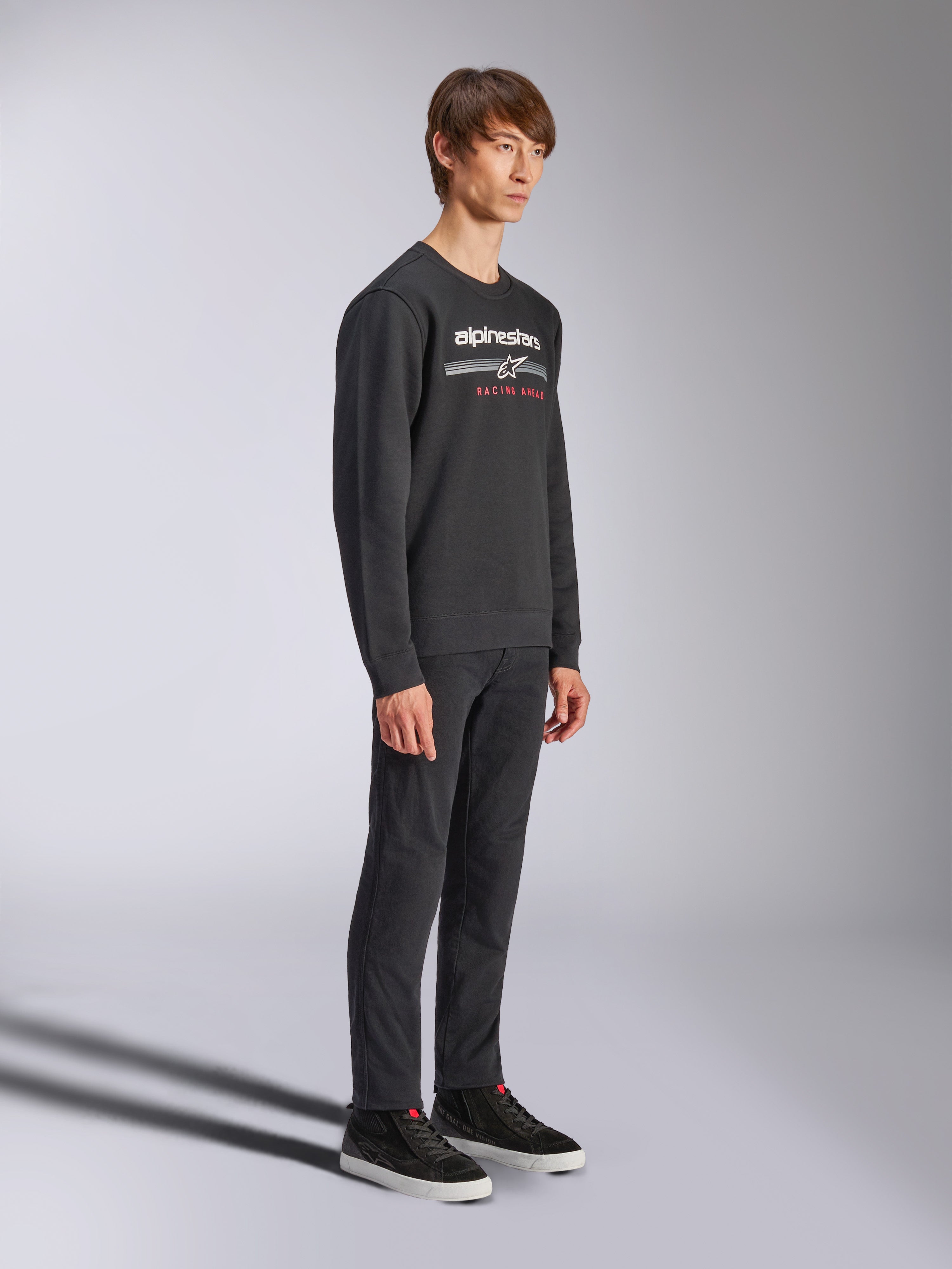 Alpinestars Bettern Crew Sweatshirt, zwarte pullover met ronde hals, wit Alpinestars-logo met 'RACING AHEAD' in het rood op de borst, gecombineerd met een zwarte broek en sneakers, casual op motorsport geïnspireerde streetwear