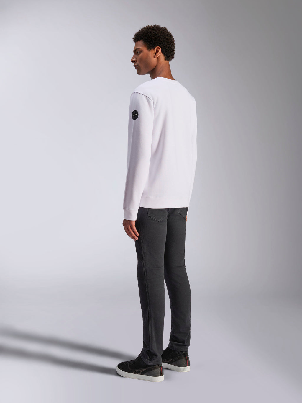 Alpinestars Bettern Crew, witte crewneck sweatshirt, achter- en zijaanzicht, zwart cirkelvormig logo op de mouw, gedragen met een donkergrijze broek en zwarte sneakers, casual motorsport lifestyle streetwear