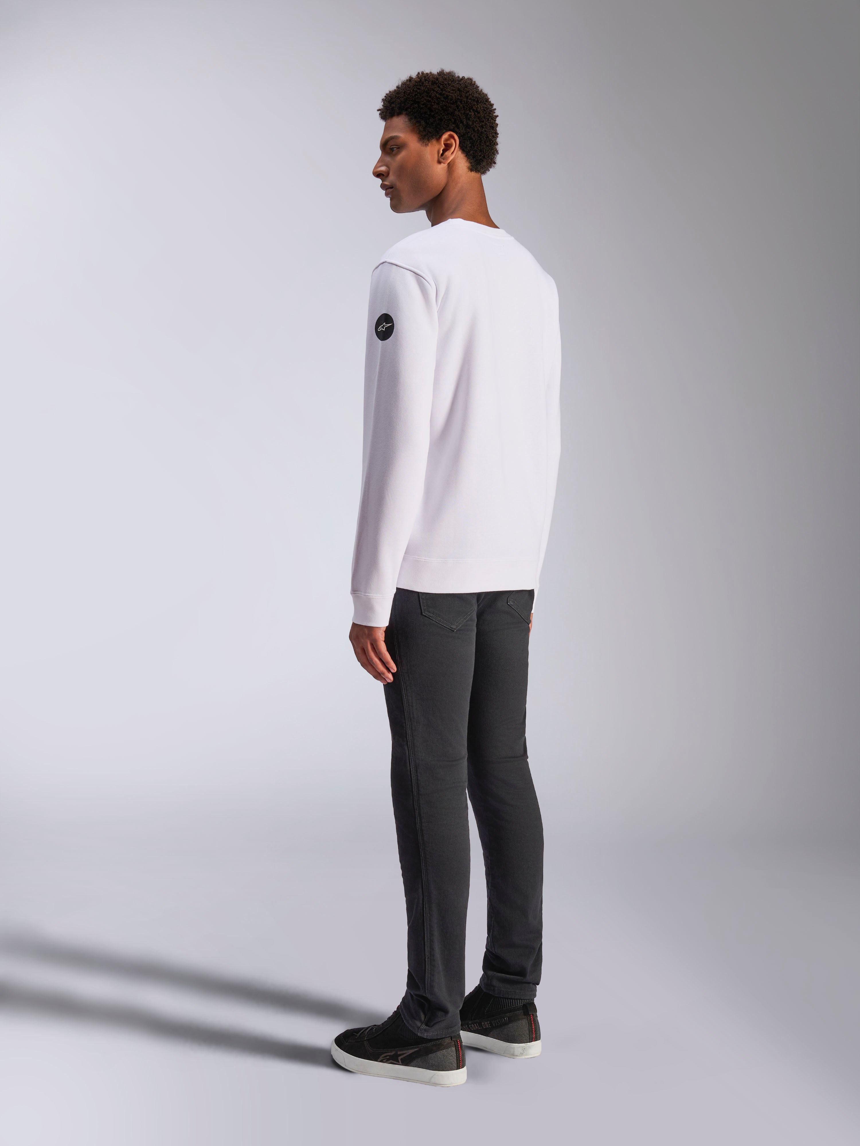 Alpinestars Bettern Crew, witte crewneck sweatshirt, achter- en zijaanzicht, zwart cirkelvormig logo op de mouw, gedragen met een donkergrijze broek en zwarte sneakers, casual motorsport lifestyle streetwear