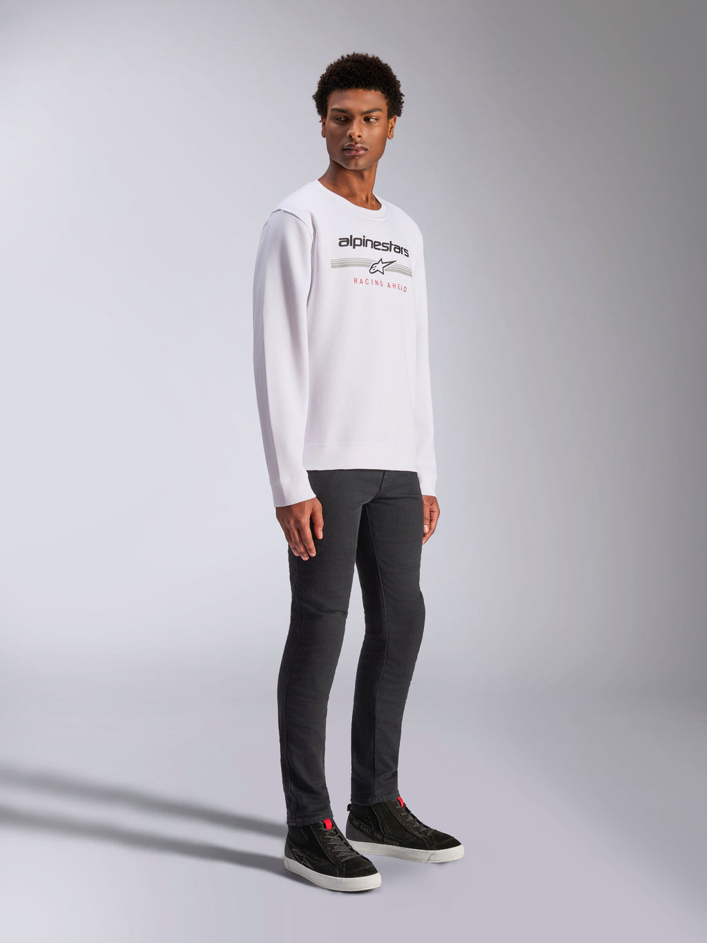 Alpinestars Bettern Crew, witte pullover sweatshirt, met zwart Alpinestars-logo en rode 'Racing Ahead'-tekst, casual motorsport streetwear, gedragen door een model met donkere broek en zwarte sneakers