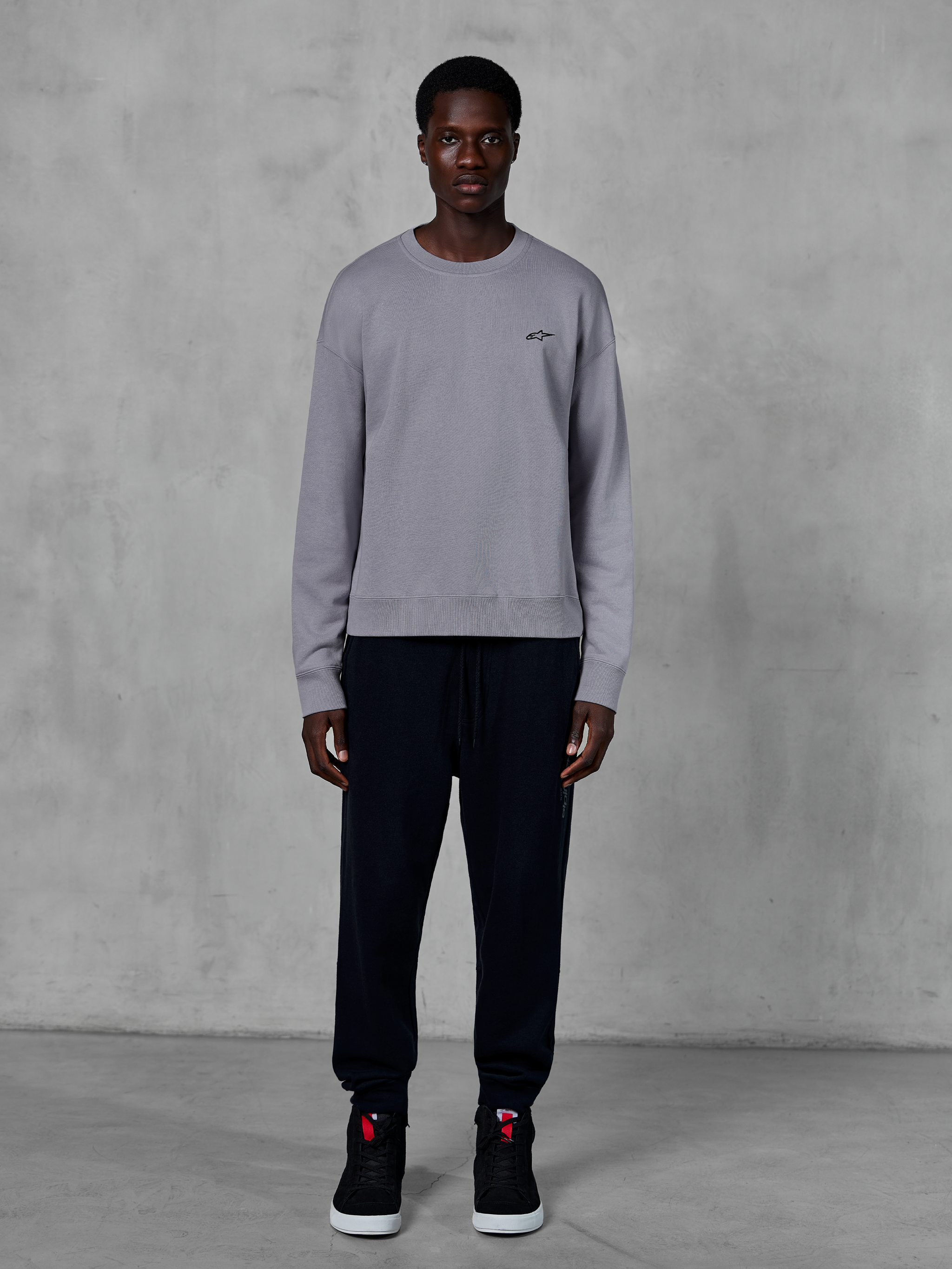 Alpinestars Essentials Ageless Crew, zilverkleurig sweatshirt met ronde hals, gedragen door een model met zwarte joggingbroek en zwarte sneakers, voorzien van een klein zwart Alpinestars-logo op de borst, casual op motorsport geïnspireerde streetwear