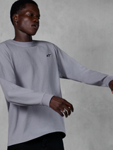 Alpinestars Essentials Ageless LS Tee, T-shirt met lange mouwen, zilver, klein zwart logo op de borst, casual op motorsport geïnspireerde streetwear, gedragen door een model tegen een grijze getextureerde achtergrond