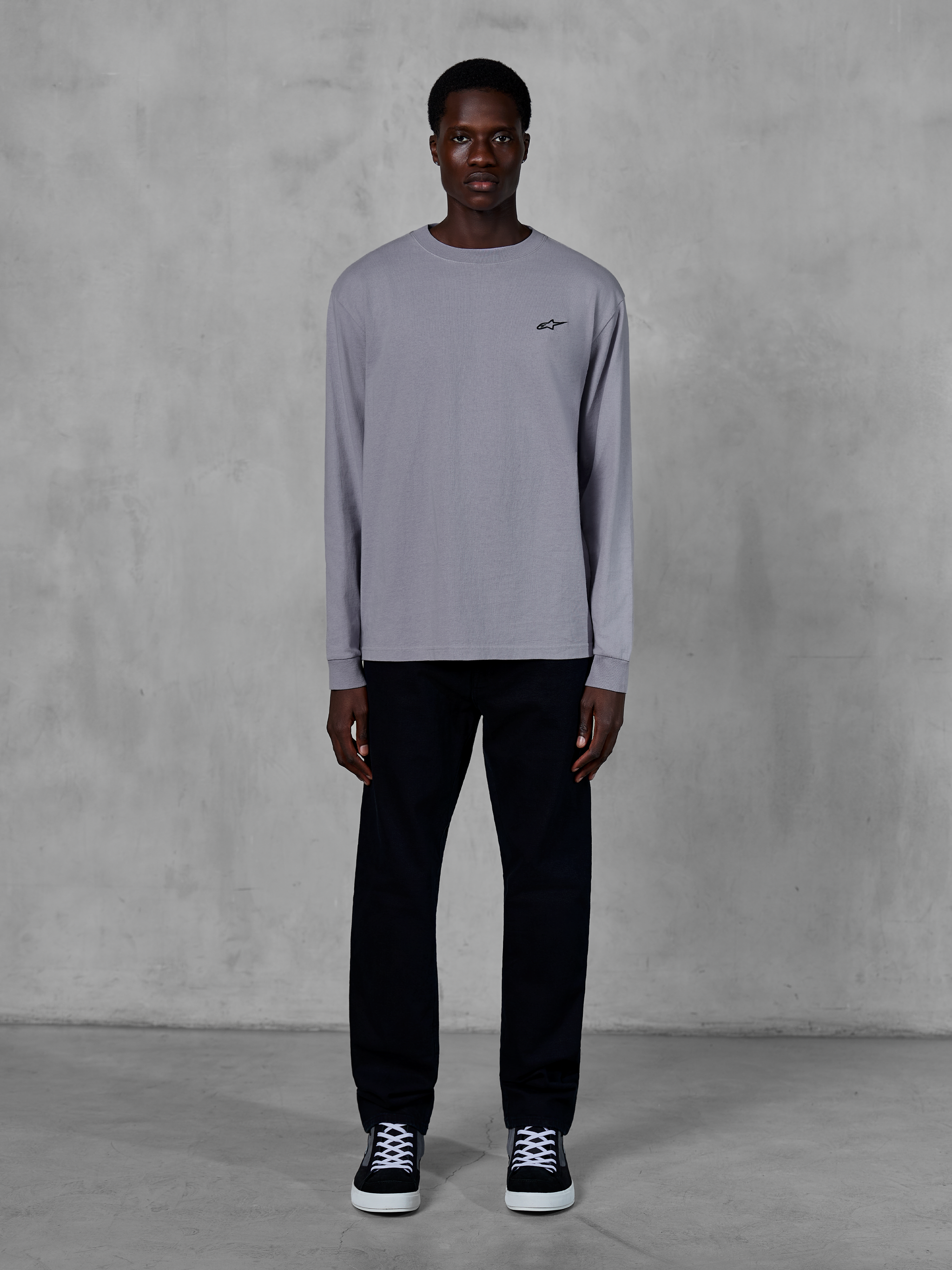 Alpinestars Essentials Ageless LS Tee, T-shirt met lange mouwen, zilver, klein zwart Alpinestars-logo op de borst, biologisch katoenen jersey met stone wash afwerking, gedragen door een model met zwarte broek en sneakers, casual streetwear