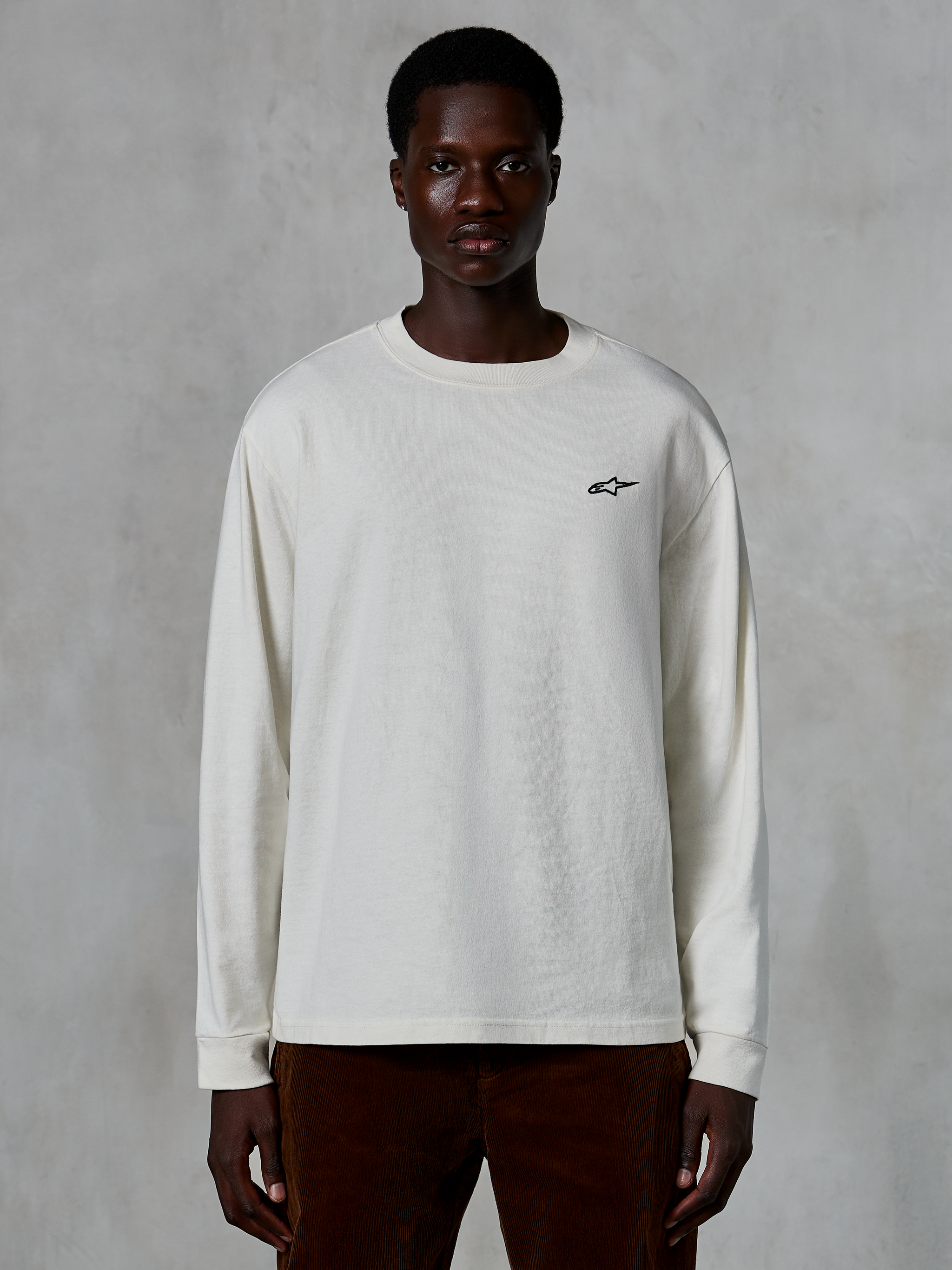 Alpinestars Essentials Ageless LS Tee, T-shirt met lange mouwen, gebroken wit, klein zwart sterlogo op de borst, casual streetwear, ontspannen pasvorm, gedragen door een model