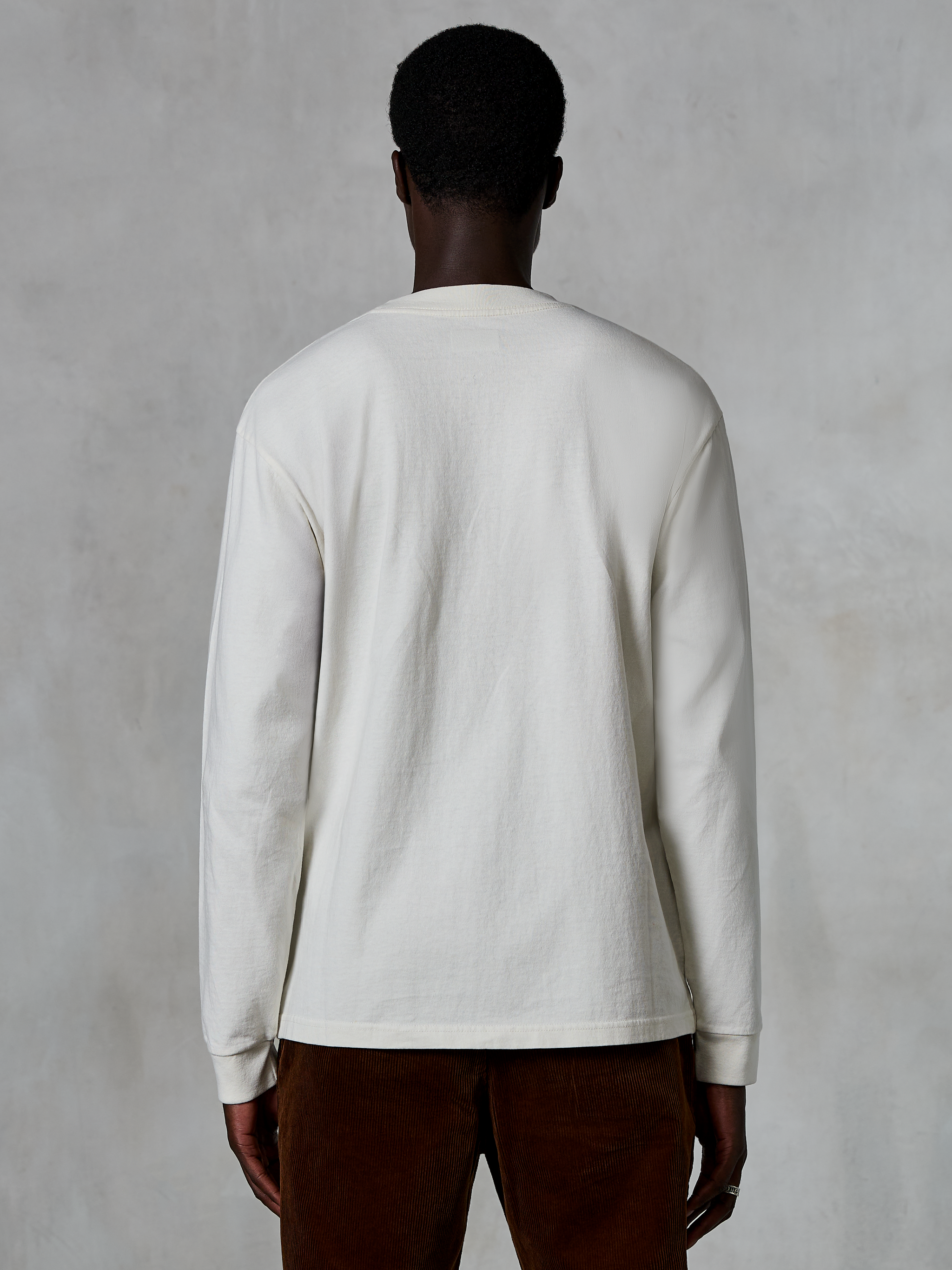 Alpinestars Essentials Ageless LS Tee, T-shirt met lange mouwen, Off White, achteraanzicht van een persoon die het shirt draagt met een bruine corduroy broek, ontspannen pasvorm, getextureerde biologische katoenen stof, minimalistische casual streetwear