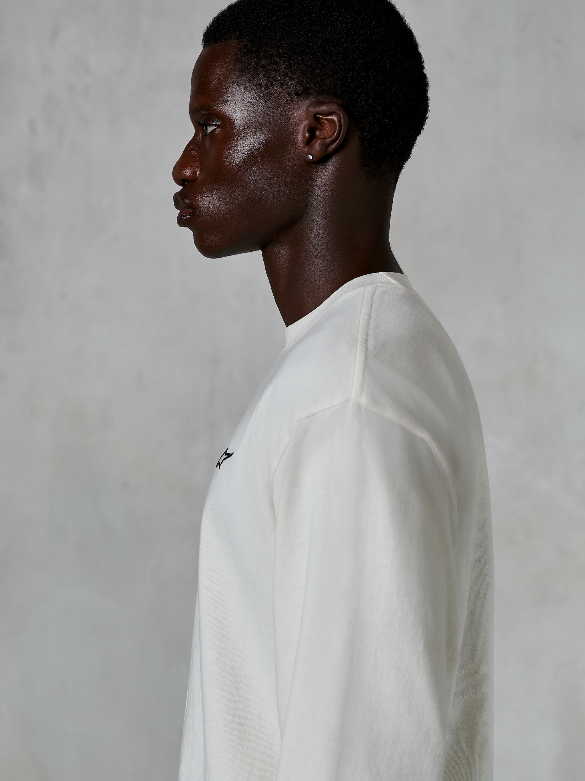 Alpinestars Essentials Ageless LS Tee, T-shirt met lange mouwen, off-white, zijaanzicht van een man die het T-shirt met relaxte pasvorm draagt, voorzien van een klein zwart logo op de borst, casual motorsport-geïnspireerde lifestyle-kleding