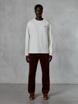 Alpinestars Essentials Ageless LS Tee, T-shirt met lange mouwen, Off-White, gedragen door model met bruine corduroy broek en bruine leren laarzen, met een klein zwart logo op de borst, ontspannen eigentijdse pasvorm, casual lifestyle streetwear