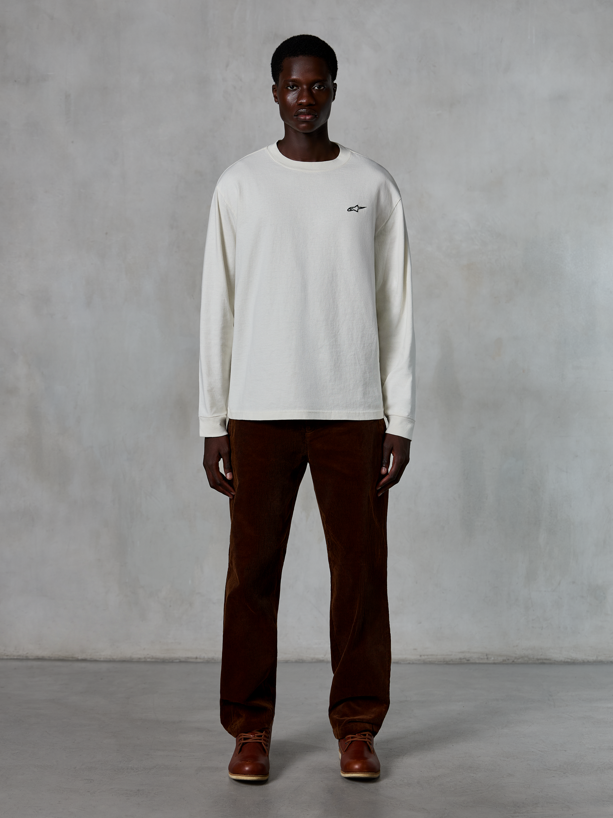 Alpinestars Essentials Ageless LS Tee, T-shirt met lange mouwen, Off-White, gedragen door model met bruine corduroy broek en bruine leren laarzen, met een klein zwart logo op de borst, ontspannen eigentijdse pasvorm, casual lifestyle streetwear