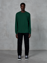 Alpinestars Essentials Ageless LS Tee, T-shirt met lange mouwen, dennengroen, voorzien van een klein zwart logo op de borst, gedragen door een model met een zwarte broek en sneakers, casual streetwear-stijl