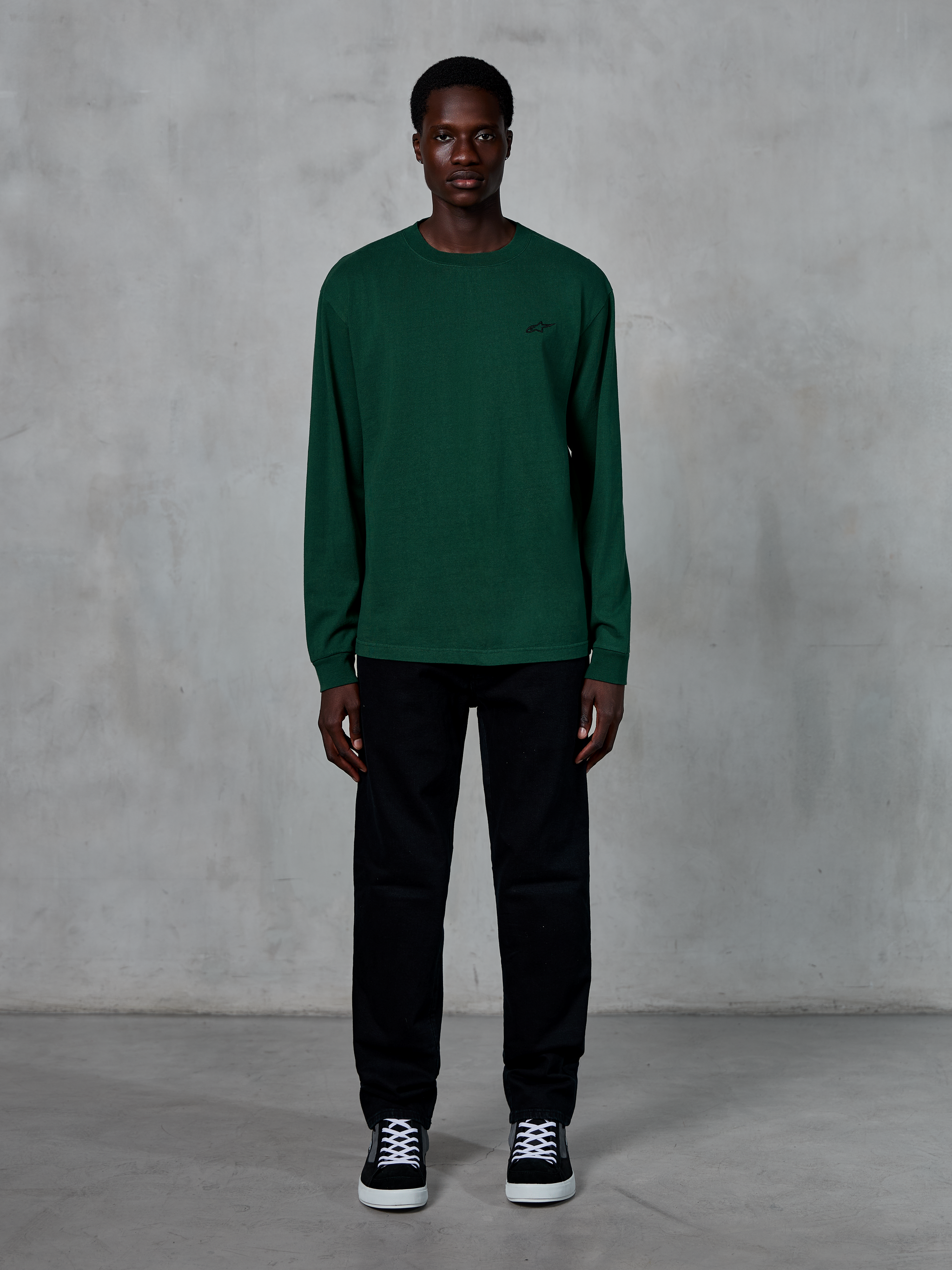 Alpinestars Essentials Ageless LS Tee, T-shirt met lange mouwen, dennengroen, voorzien van een klein zwart logo op de borst, gedragen door een model met een zwarte broek en sneakers, casual streetwear-stijl