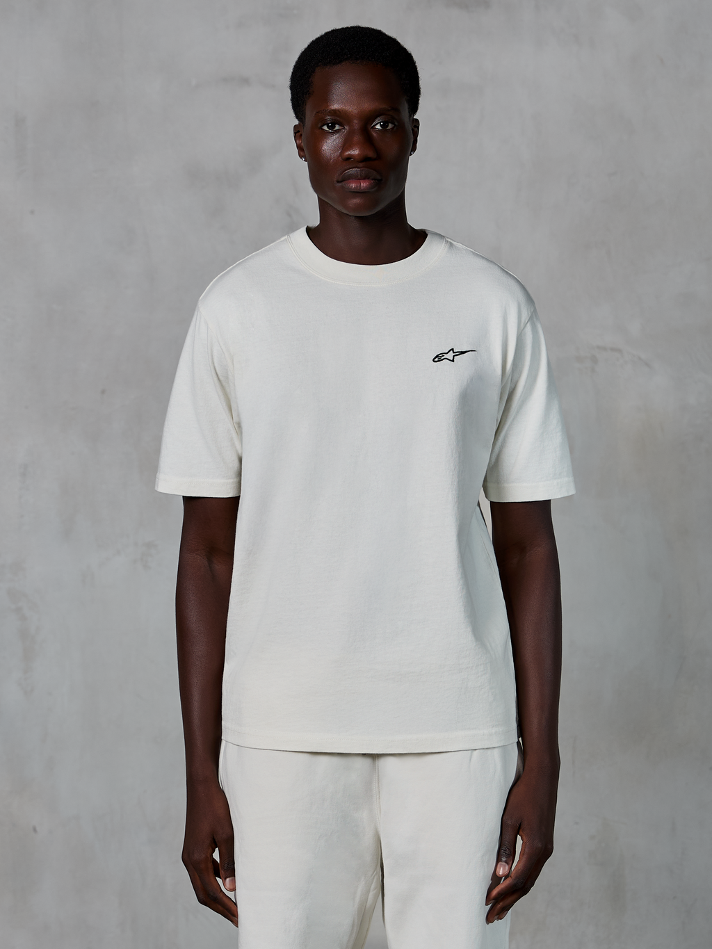 Alpinestars Essentials Ageless SS Tee, Short Sleeve T-Shirt, Off White, worn by a male model, featuring a small black Alpinestars logo on the chest, relaxed fit, organic cotton jersey, casual motorsport streetwear