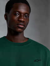 Alpinestars Essentials Ageless SS Tee, Pine Green T-shirt met korte mouwen, close-up van model in een groen biologisch katoenen shirt met een klein geborduurd logo, casual streetwear-ontwerp