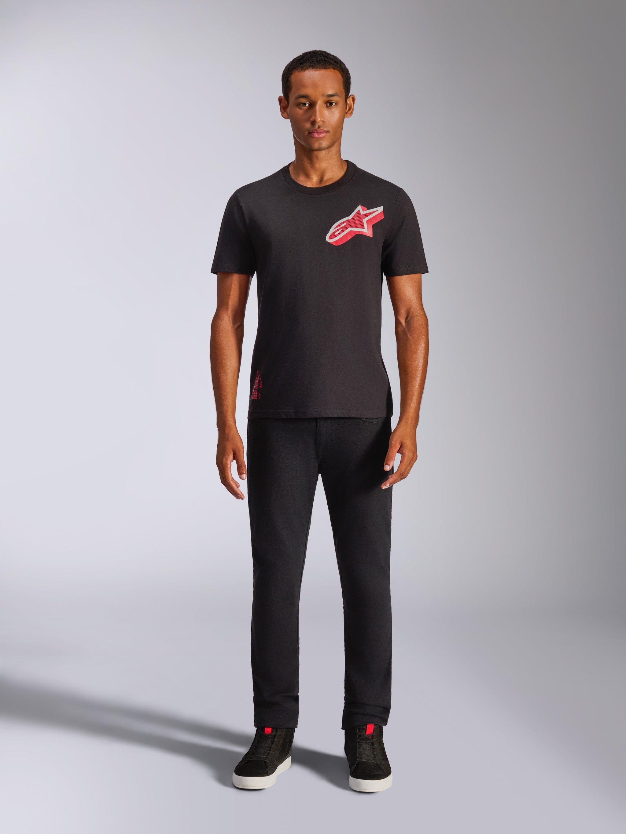 Mannelijk model draagt Alpinestars Shaded CSF Tee, zwart T-shirt met korte mouwen, met een rood schuin gradient folie Astars-logo op de borst, gecombineerd met een zwarte broek en sneakers, casual motorsport streetwear