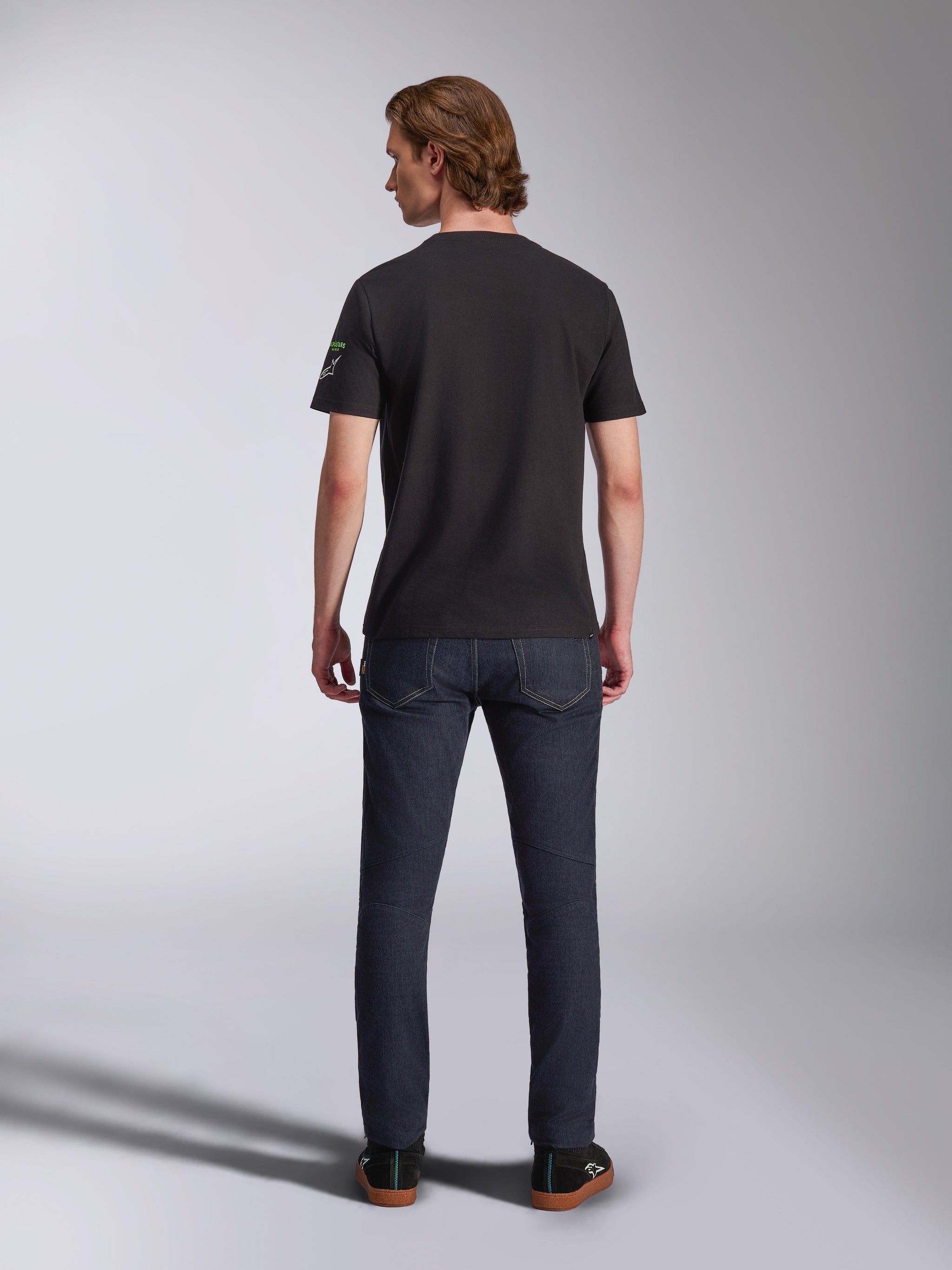 Alpinestars Entitled CSF Tee, T-shirt met korte mouwen, zwart, achteraanzicht van een mannelijk model dat het shirt draagt met een donkerblauwe denim jeans, met een klein wit en groen logo op de linkermouw, casual motorsport-geïnspireerde streetwear