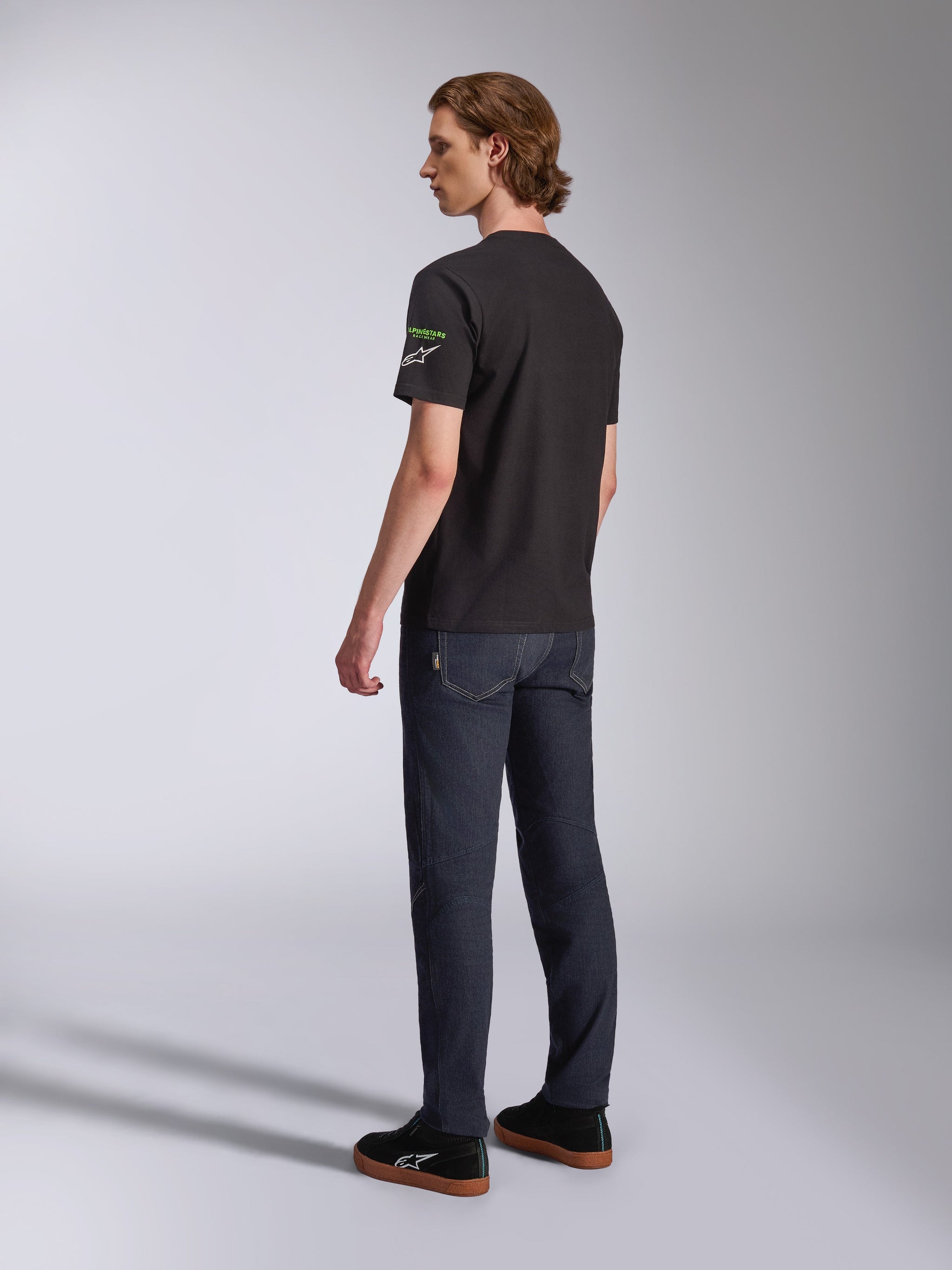 Alpinestars Entitled CSF Tee, T-shirt met korte mouwen, zwart, achteraanzicht in profiel van een model dat het T-shirt draagt met groene en witte logo's op de mouw, gecombineerd met een donkere spijkerbroek en zwarte sneakers, casual motorsport streetwear