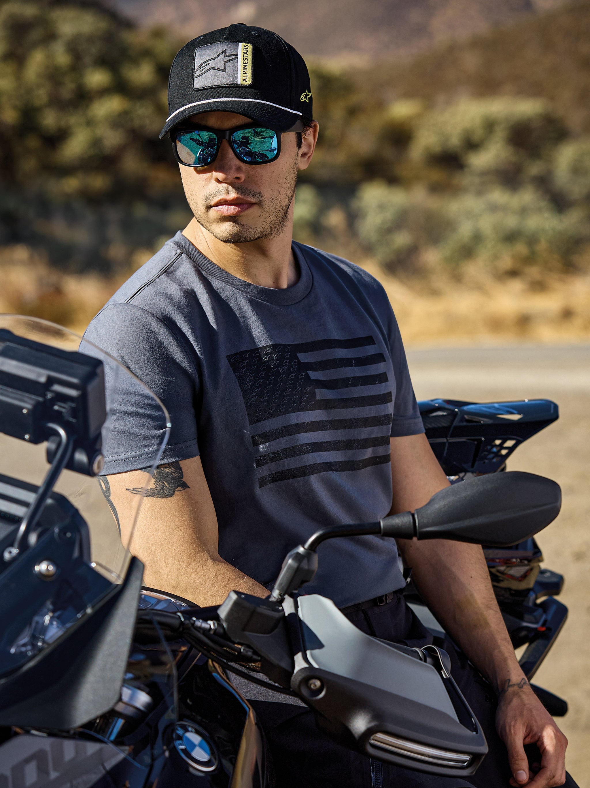 Alpinestars Waving CSF Tee, Short Sleeve T-Shirt, Charcoal, featuring a black waving flag graphic on the chest, worn by a man sitting on a motorcycle in a sunny outdoor setting, casual motorsport lifestyle