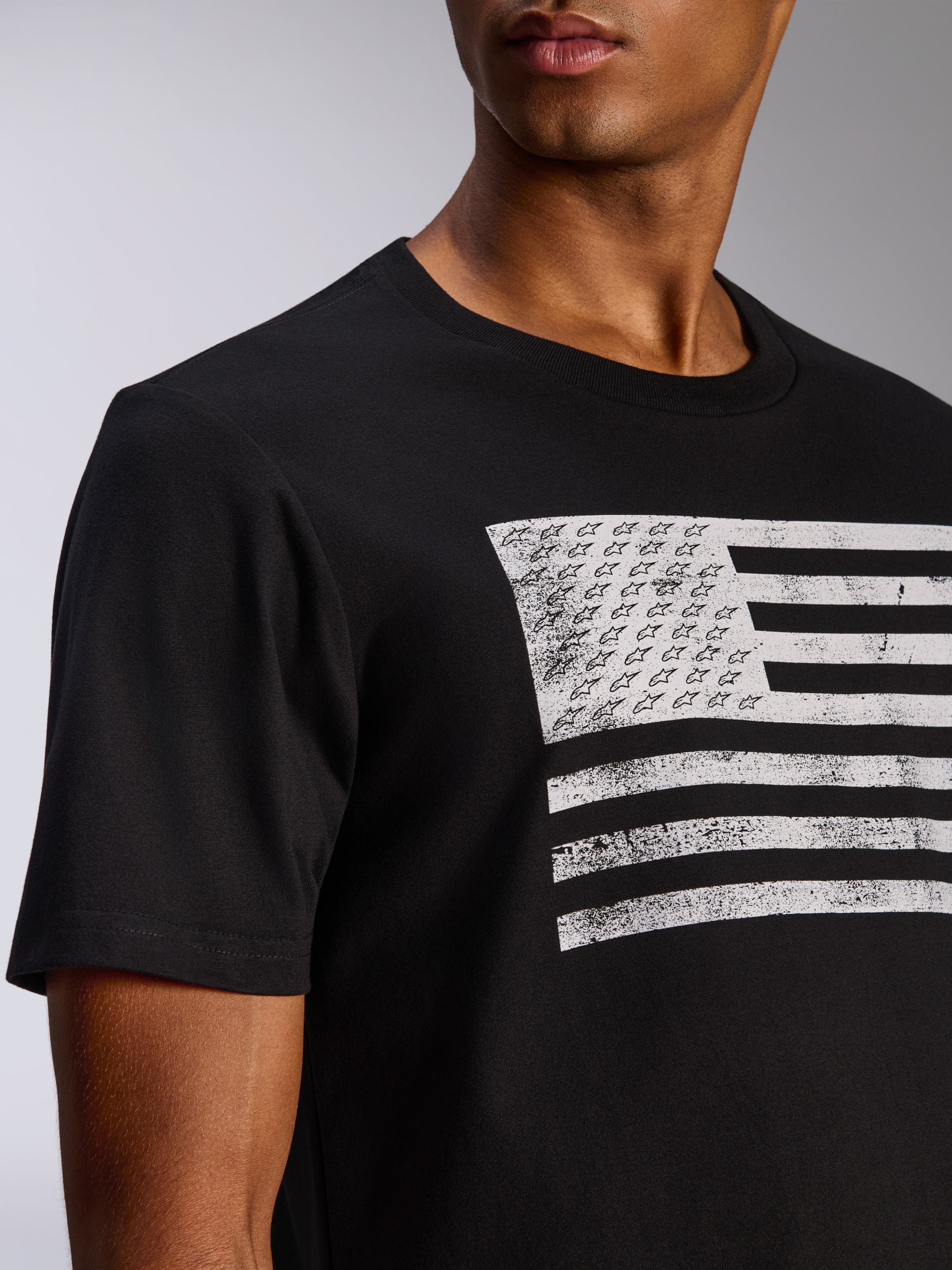 Alpinestars Waving CSF Tee, T-shirt met korte mouwen, zwart, verweerde witte vlag-grafische print met Alpinestars sterren en strepen, casual motorsportkleding, close-up van een persoon die het shirt draagt
