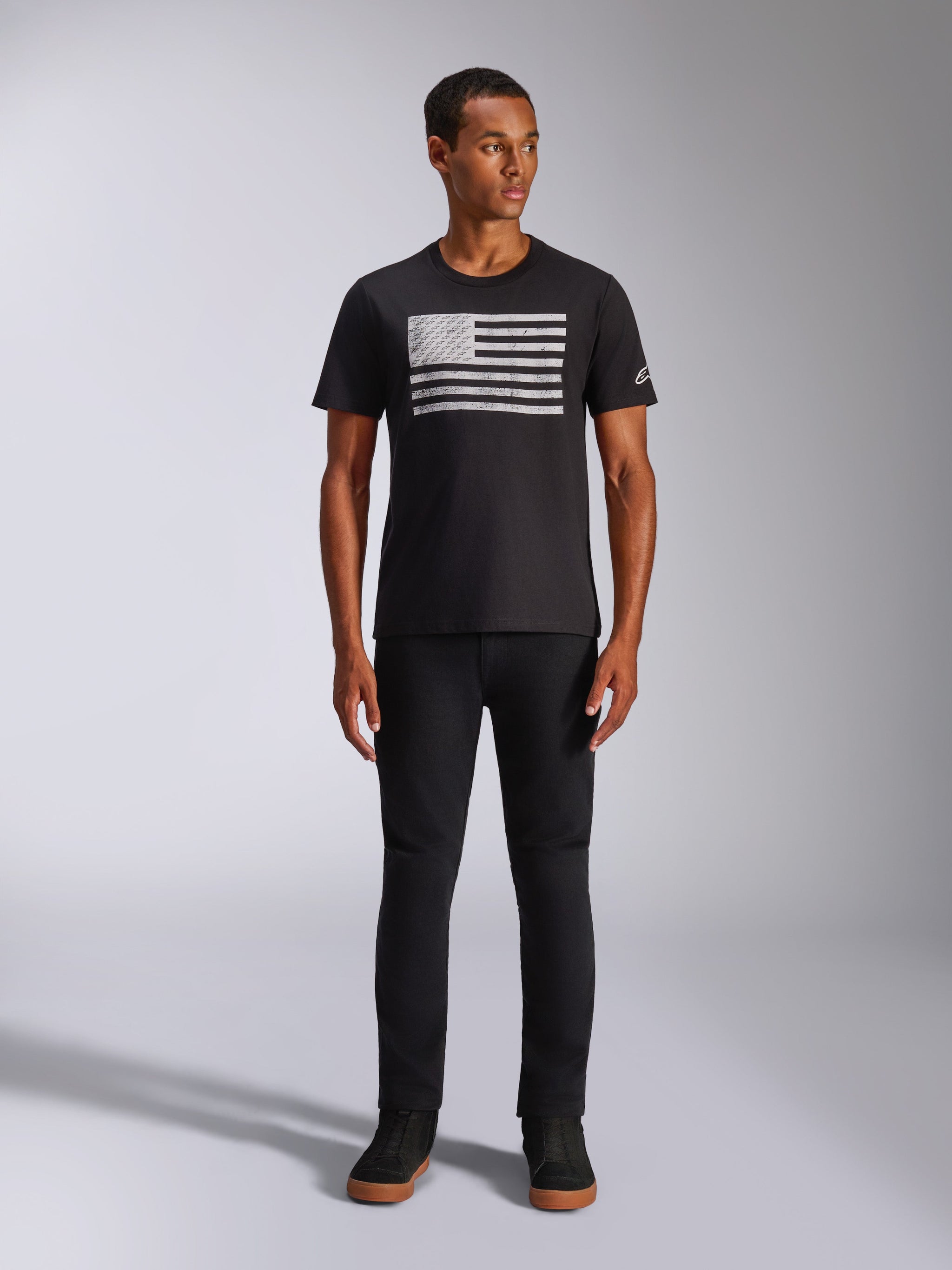 Alpinestars Waving CSF Tee, T-shirt met korte mouwen, zwart, met een verweerde witte graphic in vlagstijl op de borst, gedragen door een model met zwarte broek en zwarte hoge sneakers, casual motorsport streetwear