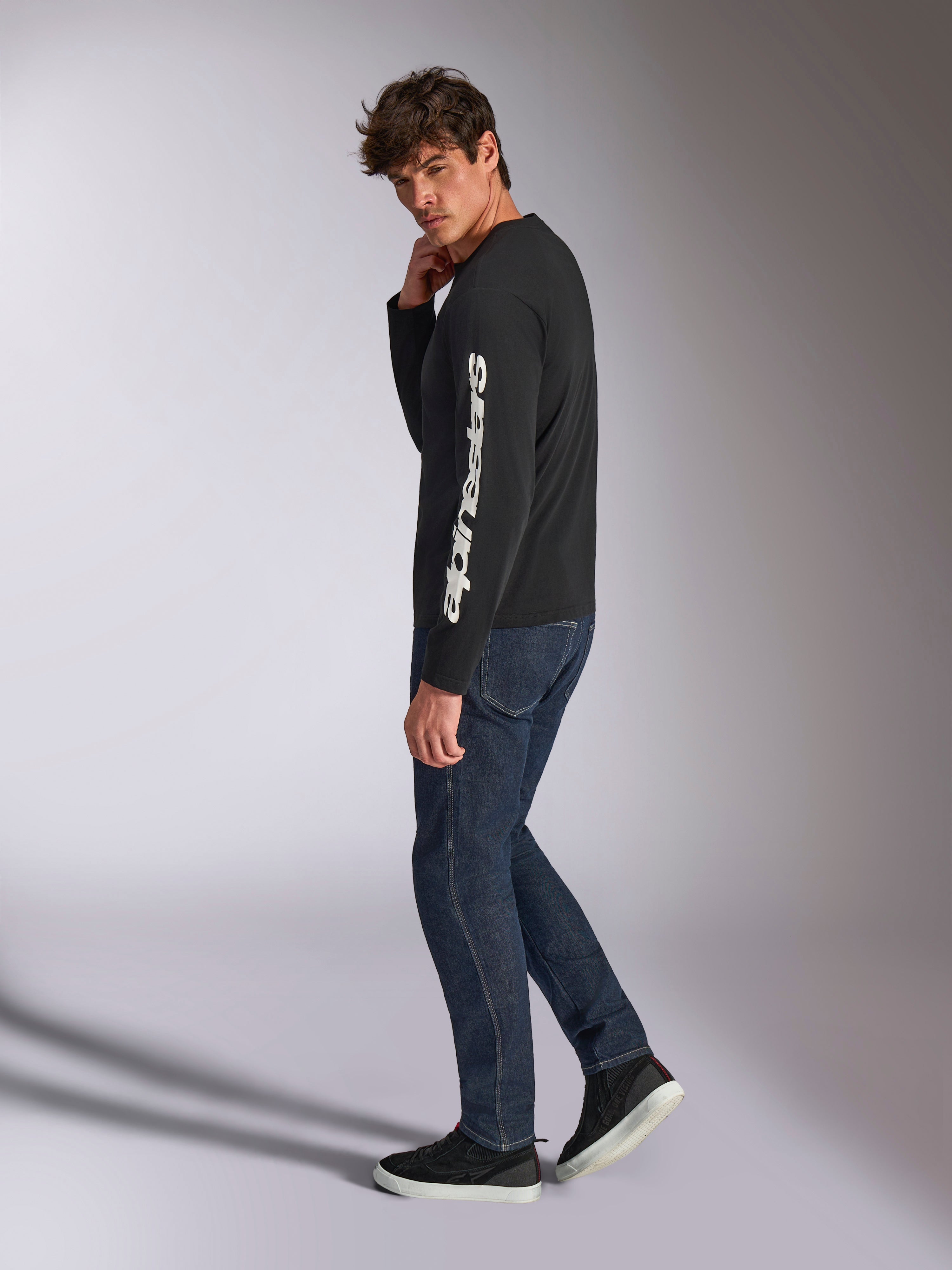 Alpinestars Trackside CSF Long Sleeve Tee, Black with white vertical Alpinestars wordmark on the sleeve, worn by a model with dark denim and sneakers, casual motorsport-inspired sportswear