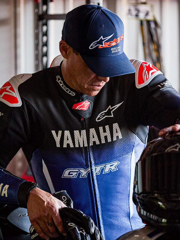 Alpinestars Entitled pet, marineblauw met oranje Astars-logo en witte 'Racing Ahead'-tekst, gedragen door een rijder in een blauw en zwart Yamaha-racipak, baseballcap met gebogen klep en gestructureerde kroon