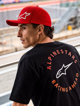 Alpinestars Corp Shift Multi Hat, Red curved bill cap with white 3D Astars logo, worn with a black Alpinestars t-shirt featuring 'Racing Ahead' graphic, lifestyle motorsport setting at a racing circuit
