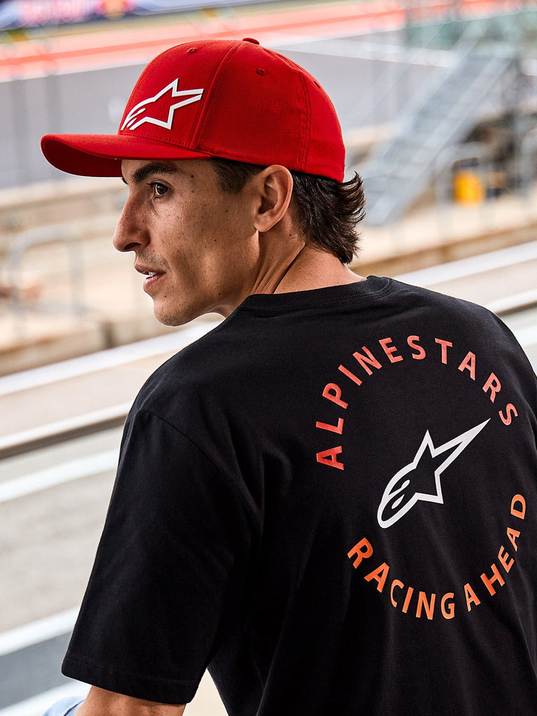 Alpinestars Corp Shift Multi Pet, rode pet met gebogen klep en wit 3D Astars-logo, gedragen met een zwart Alpinestars t-shirt met 'Racing Ahead' graphic, lifestyle motorsport setting op een racecircuit