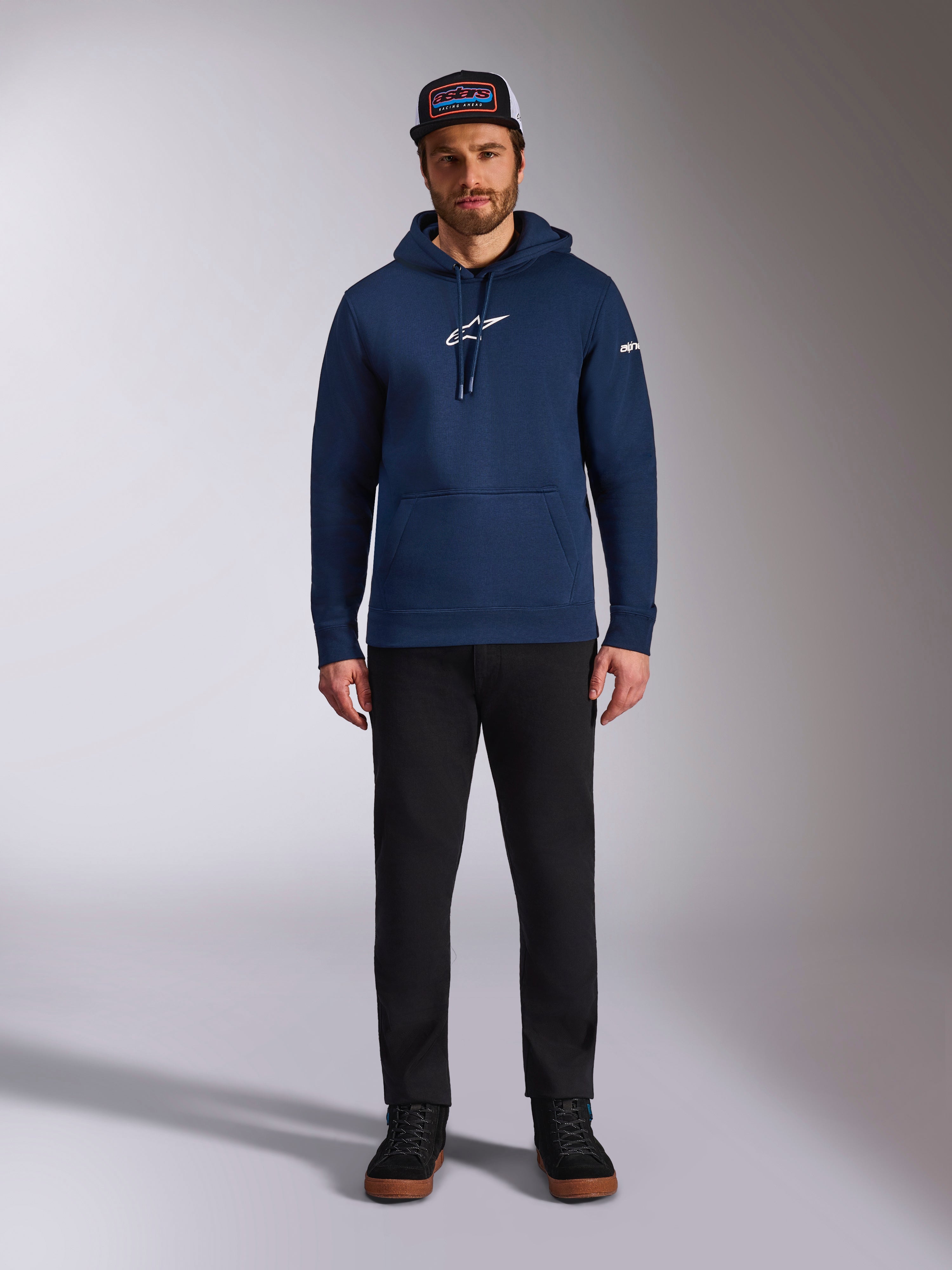 Male model wearing the Alpinestars Frontal Hoodie in Navy, pullover fleece with a white Astars logo on the chest and word logo on the sleeve, featuring a kangaroo pocket and drawstring hood, paired with black pants and a branded cap, casual motorsport streetwear