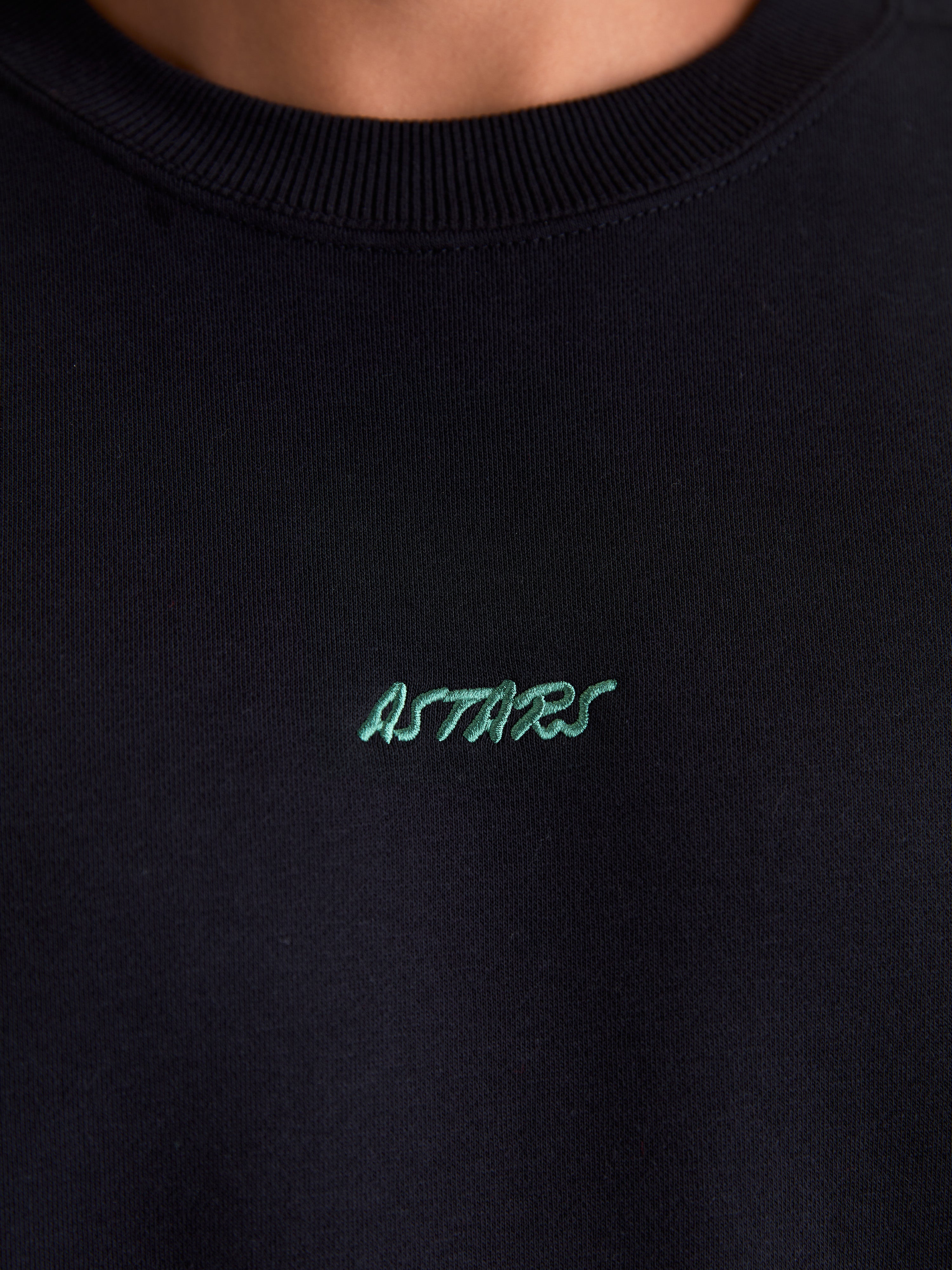 Alpinestars Guise Oversized Crew, zwarte sportswear fleece pullover, detailopname van de geribde ronde hals en het groene geborduurde Astars-logo op de borst, gemaakt van een zachte mix van katoen en gerecycled polyester.