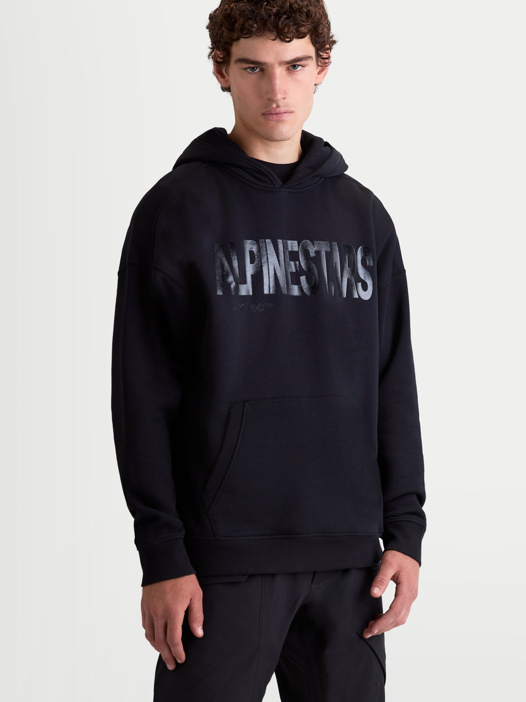 Alpinestars Shaw Oversized Hoodie, ontspannen sportswear-pullover voor dagelijks comfort, vooraanzicht op model, zwart, voorzien van een getextureerd logo op de borst, kangoeroezak en zelfgevoerde capuchon, gemaakt van een katoen en gerecycled polyester fleece mix.