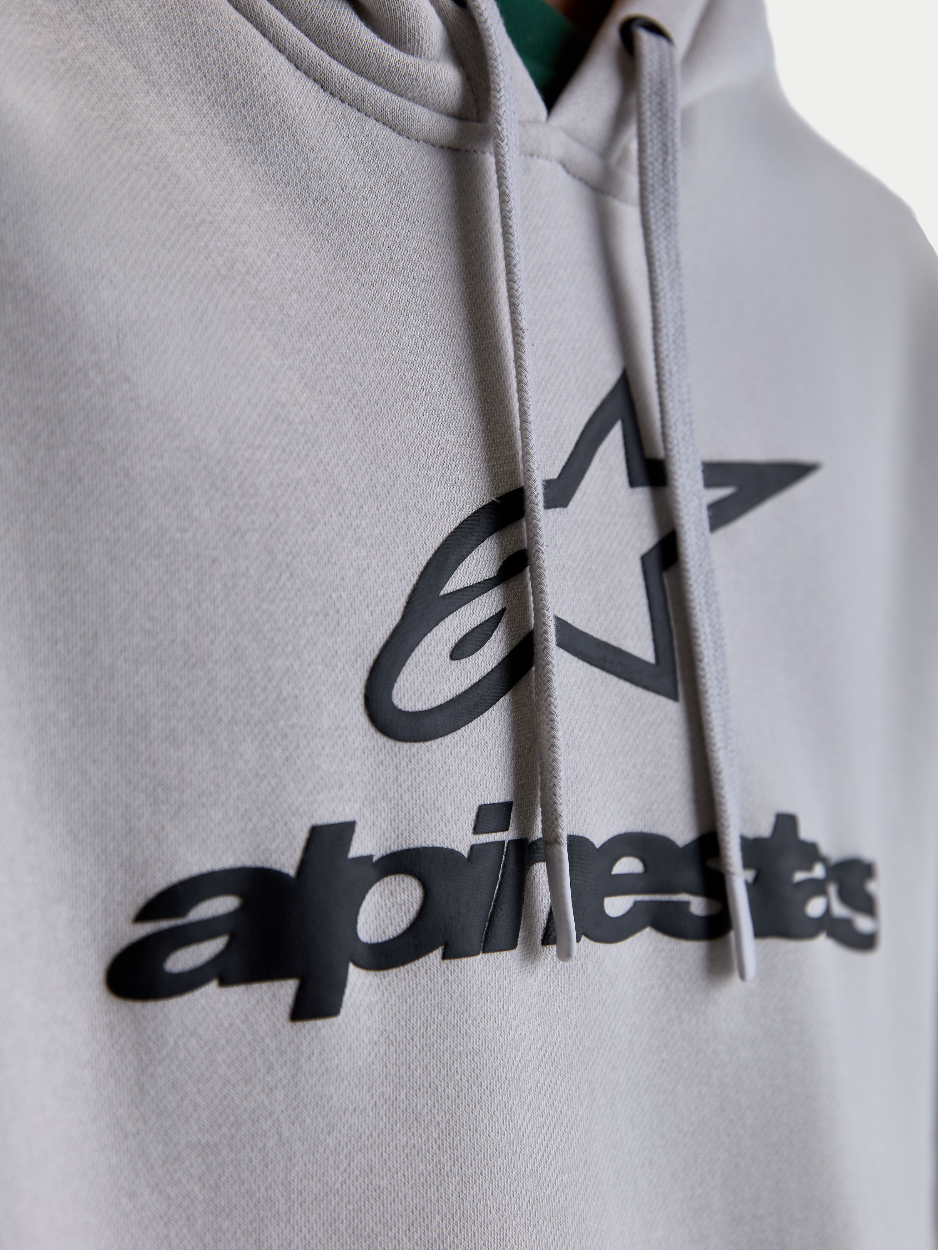 Alpinestars Prelude oversized hoodie, close-up detail van het zwarte puff-print logo en de ster-graphic op cementgrijs fleece, voorzien van platte trekkoorden, premium unisex lifestyle sportswear hoodie.