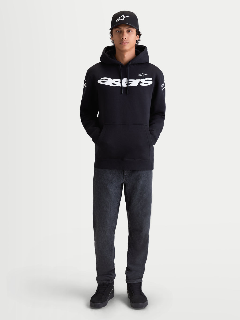 Alpinestars Velocifier hoodie, heren-fleece pullover voor casual gebruik, zwart met wit Astars-woordmerk op de borst en logo's op de schouders, premium katoenmix, getoond op model met bijpassende pet.