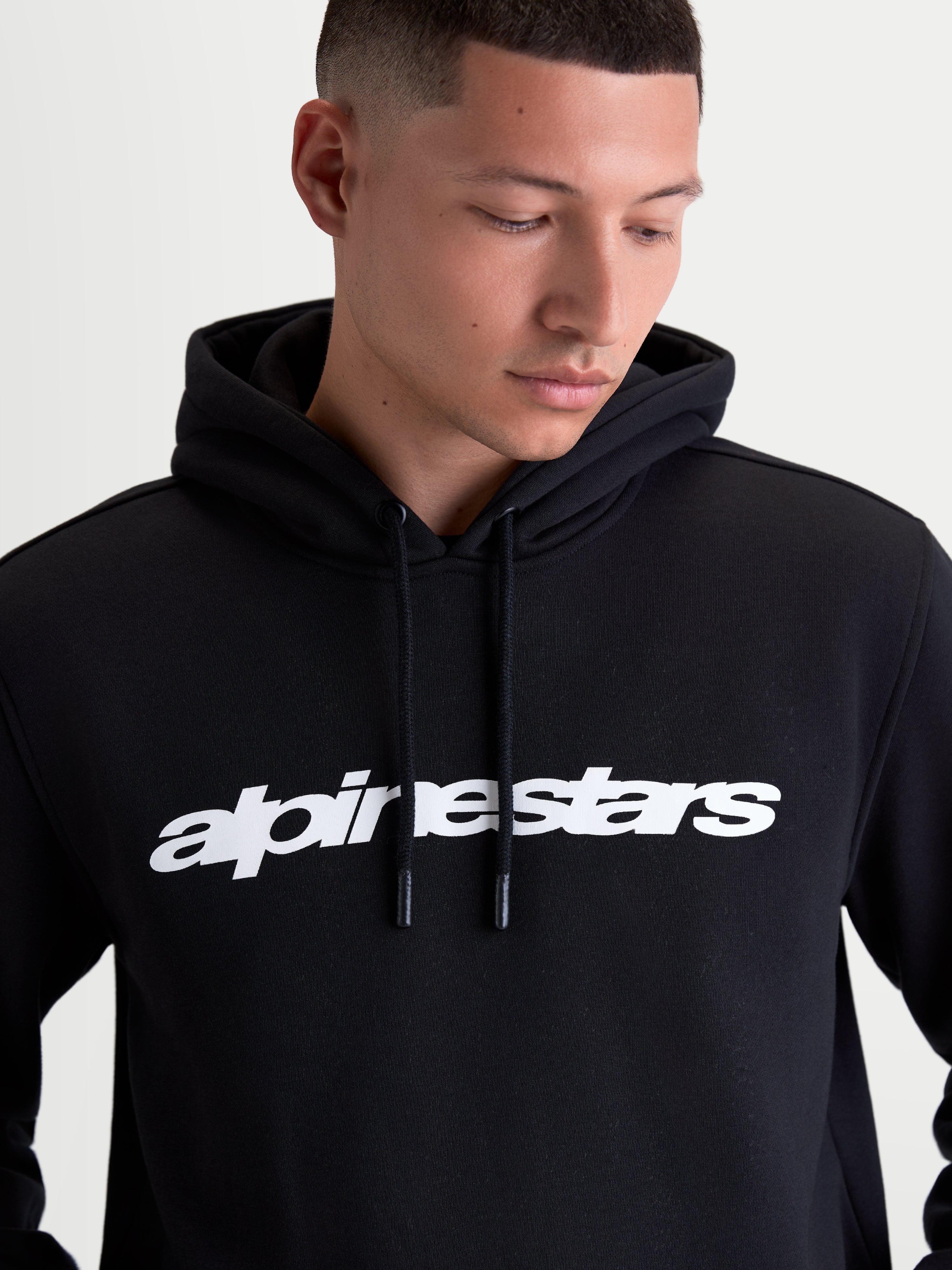 Alpinestars Race Wordmark Hoodie gedragen door een model, zwarte sportswear-hoodie met een opvallend wit woordmerklogo op de borst, gevoerde capuchon en trekkoorden met coating, gemaakt van premium katoen en gerecycled polyester fleece.