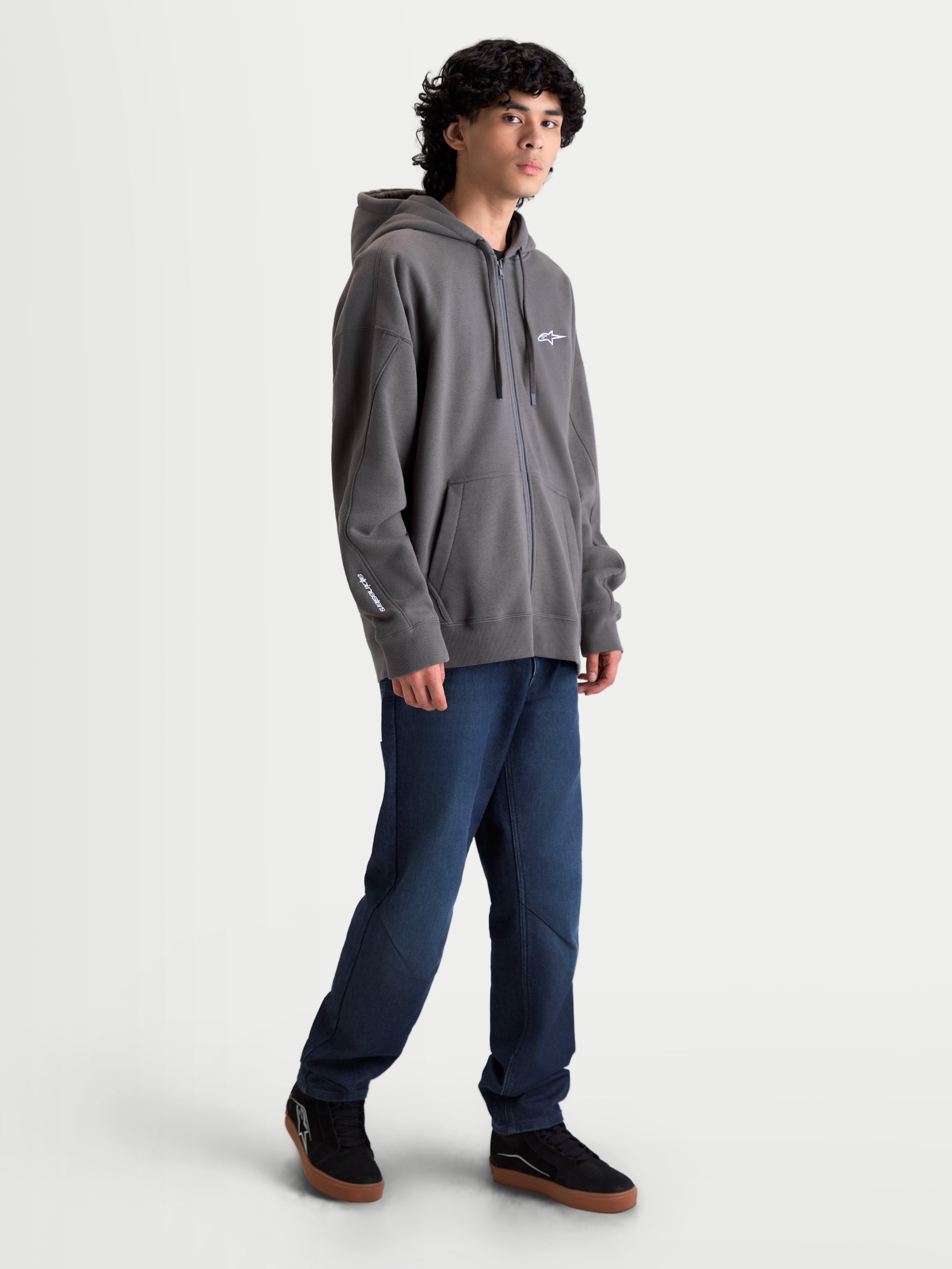 Alpinestars Entity oversized hoodie, sportswear fleece hoodie met ritssluiting, grijs, voorzien van een oversized pasvorm, Vislon-rits aan de voorzijde, kangoeroezakken en wit geborduurd Alpinestars-logo op de borst en mouw. Casual motorsportgeïnspireerde lifestyle kleding gedragen door een model.
