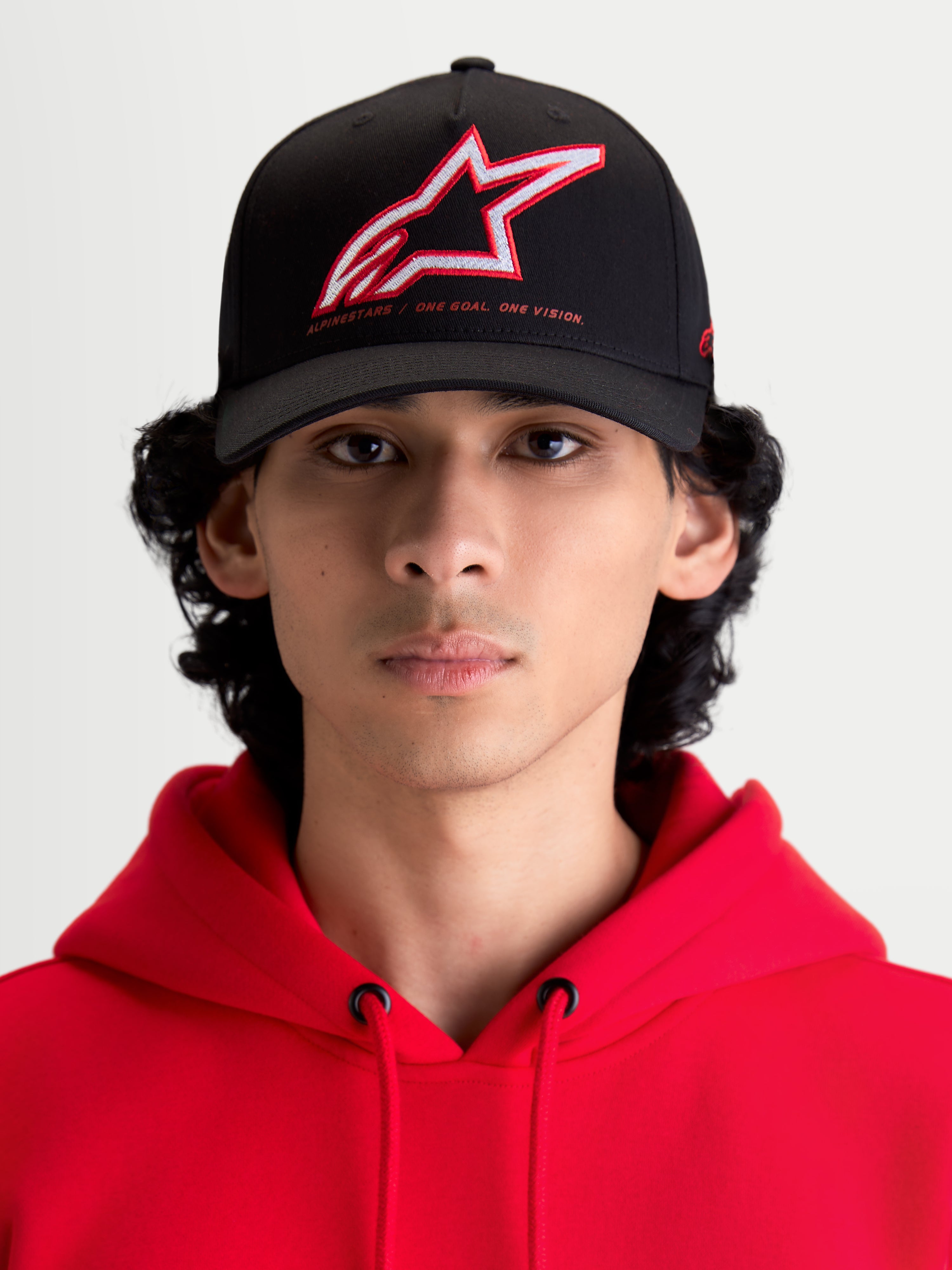 Alpinestars Lineout pet, sportswear baseballpet met gebogen klep, zwart met rood en wit geborduurd Astars-logo en One Goal One Vision-branding, vooraanzicht van model met een rode hoodie, gestructureerde keperstofconstructie.