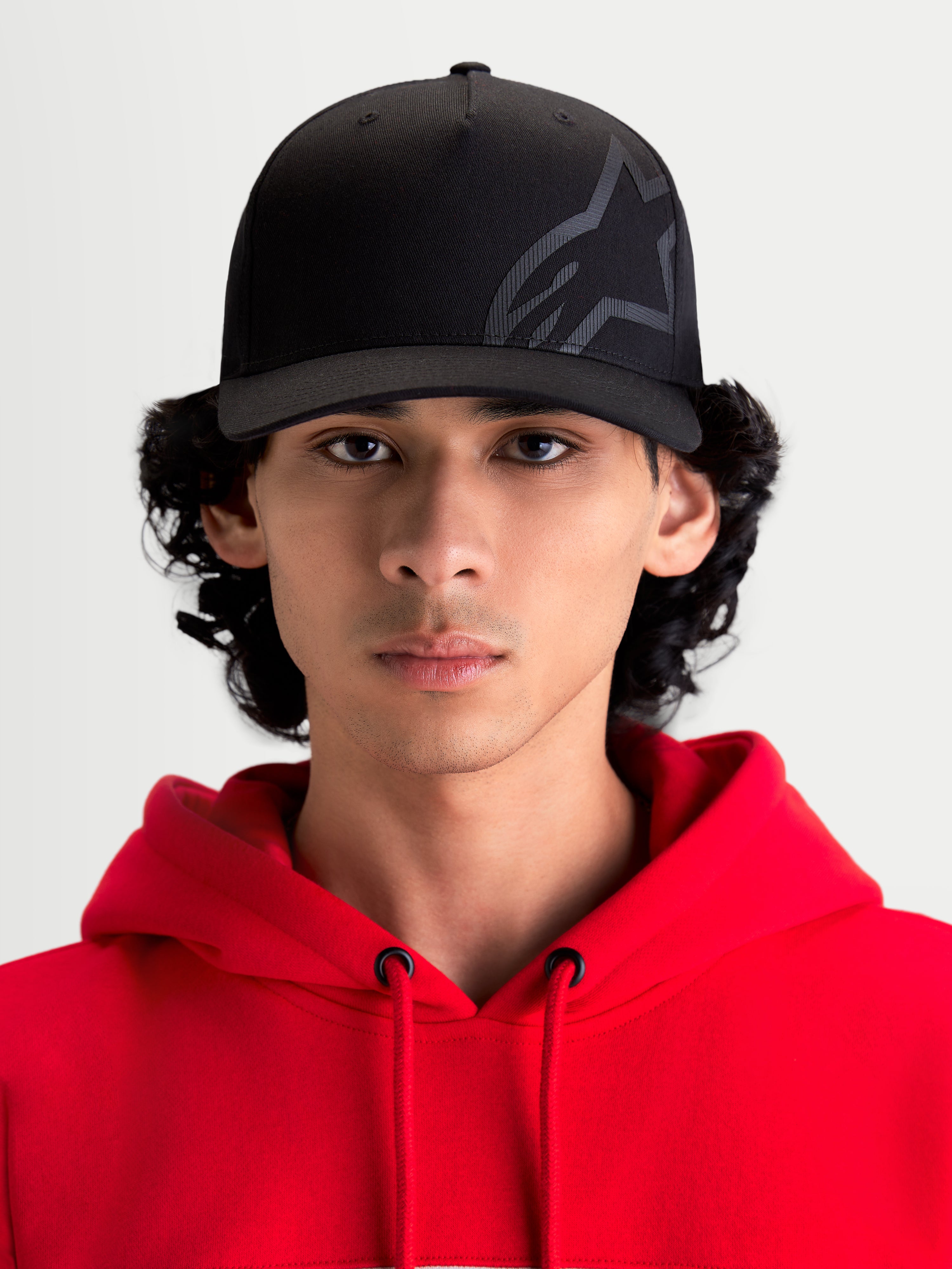 Alpinestars Maze pet, zwarte motorsport-geïnspireerde sportswearpet, vooraanzicht op model met rode hoodie. Voorzien van gestructureerde kroon, gebogen klep en getextureerd Astars Corp Shift-logo op duurzame keperstof voor casual stijl naast de motor.