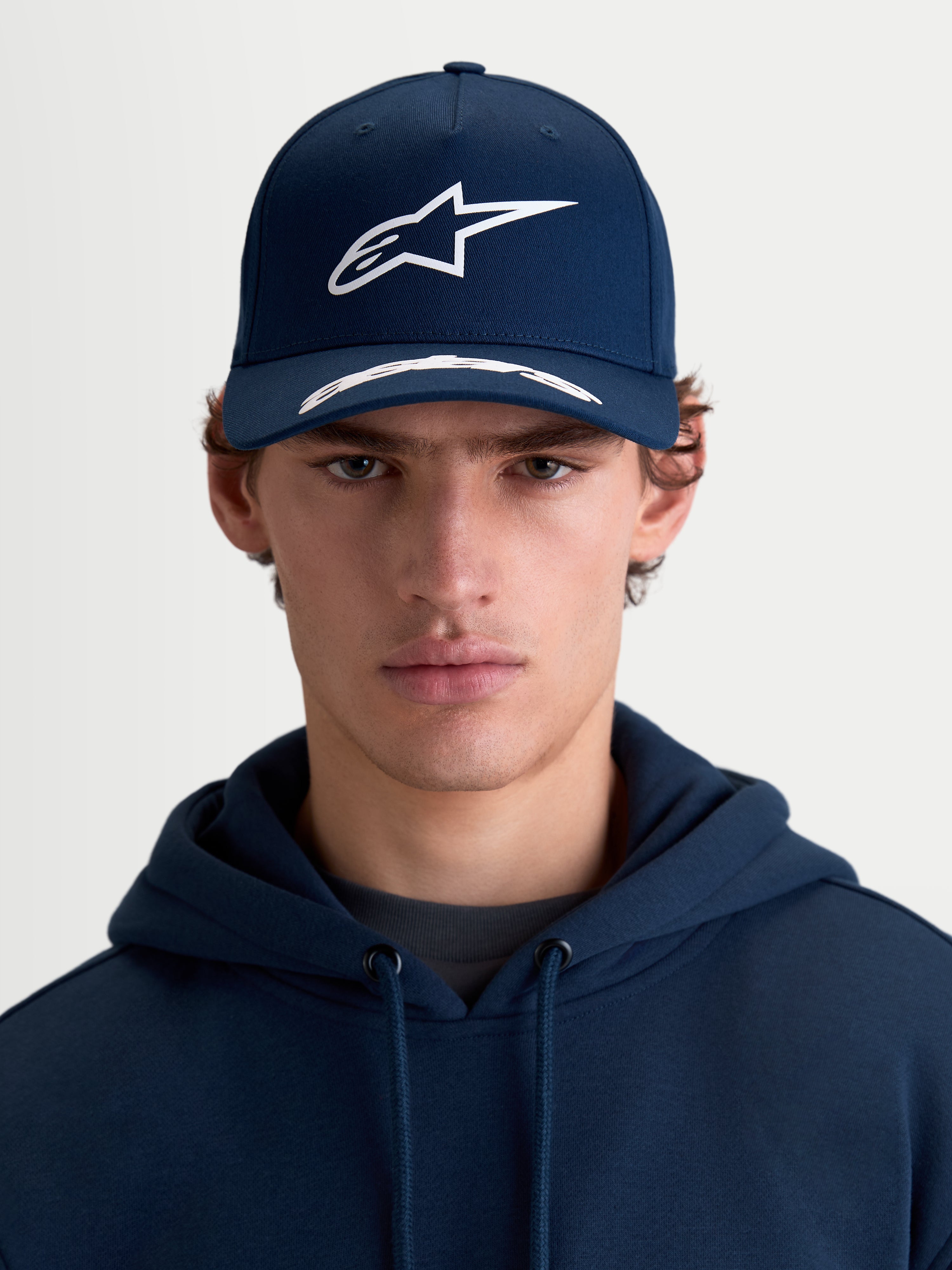 Alpinestars Arclight pet, sportswear-pet met gebogen klep, donkerblauw en wit, vooraanzicht op model, voorzien van een gestructureerde kroon, getextureerd puffprint Astars-logo en premium katoenen keperstof.
