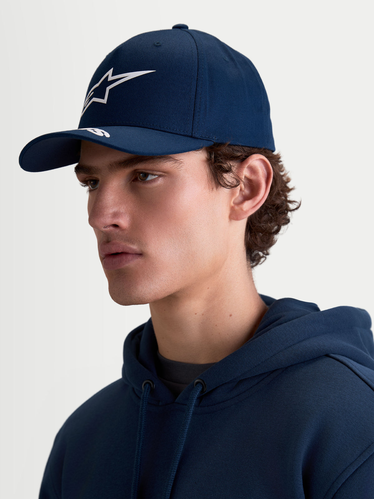 Alpinestars Arclight pet, donkerblauwe sportswear baseballpet voor heren en dames, met een gebogen klep, witte puffprint Astars-branding en gestructureerde kroon. Zijaanzicht op model toont de klassieke motorsportpasvorm en duurzame katoenen keperstof.