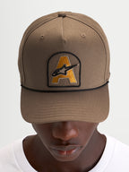 Alpinestars Acme Snapback Pet, gebogen klep cap voor dagelijkse sportswear, walnootbruin met Astars-logoborduursel patch en zwart koorddetail, gedragen door een model