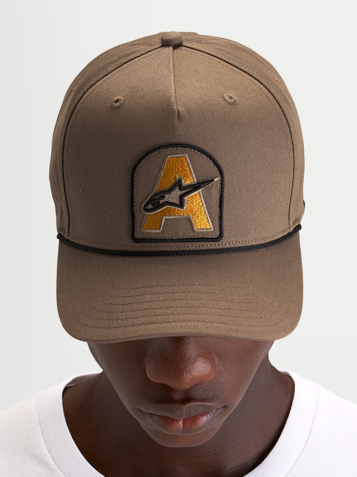 Alpinestars Acme Snapback Pet, gebogen klep cap voor dagelijkse sportswear, walnootbruin met Astars-logoborduursel patch en zwart koorddetail, gedragen door een model