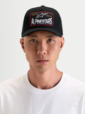Alpinestars Hemmings Trucker pet, zwarte sportswear pet met gebogen klep en ademende mesh achterkant, met een geweven Alpinestars-logopatch op de voorkant, gedragen door een model