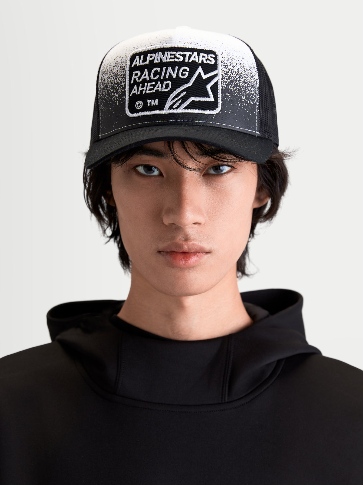 Alpinestars Gravel Trucker pet, sportswear lifestyle-pet, zwart en wit, vooraanzicht op model, gebogen klep, ademende mesh achterkant en Racing Ahead geweven patch met grindtextuurdetails.