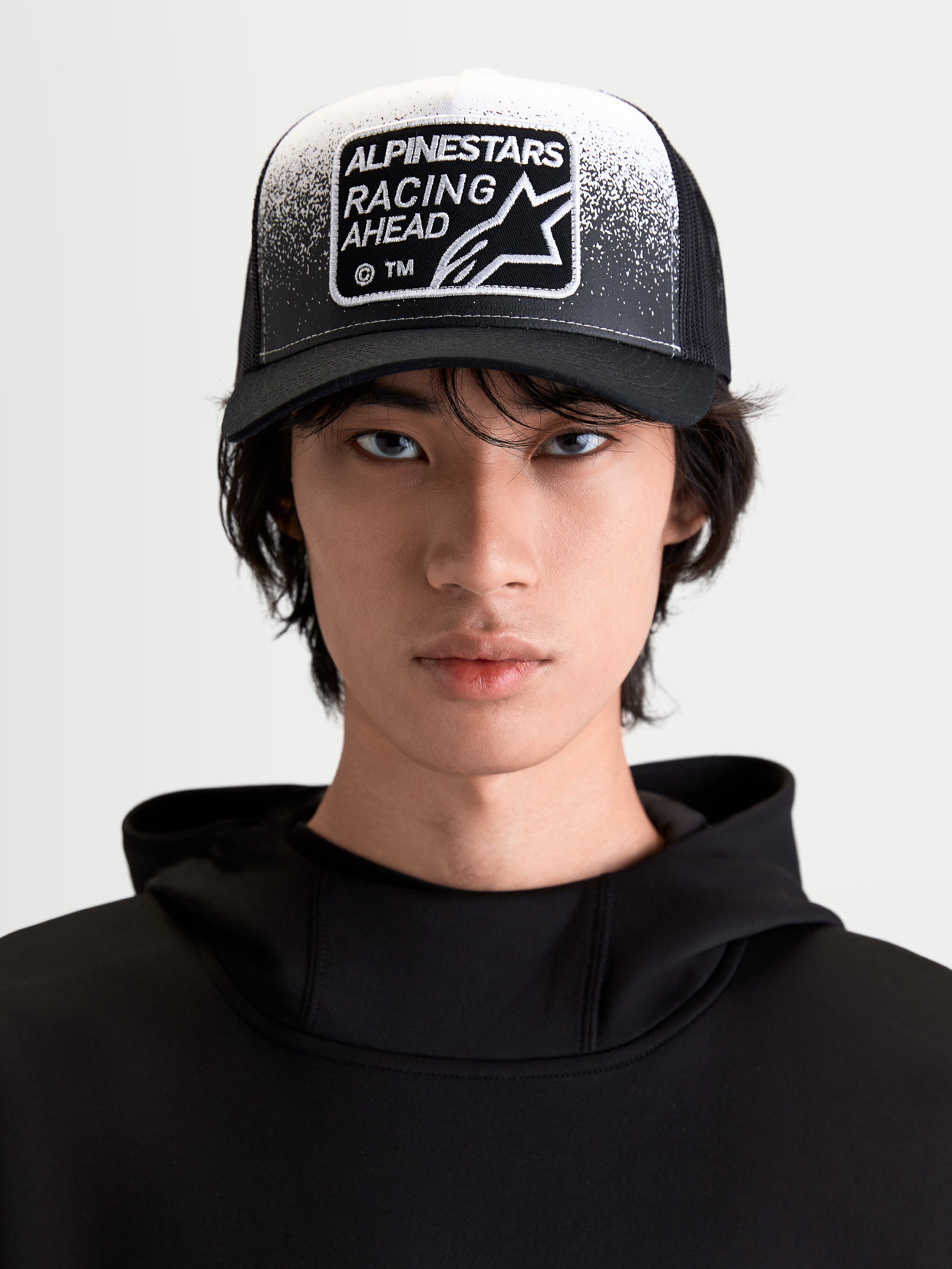 Alpinestars Gravel Trucker pet, sportswear lifestyle-pet, zwart en wit, vooraanzicht op model, gebogen klep, ademende mesh achterkant en Racing Ahead geweven patch met grindtextuurdetails.